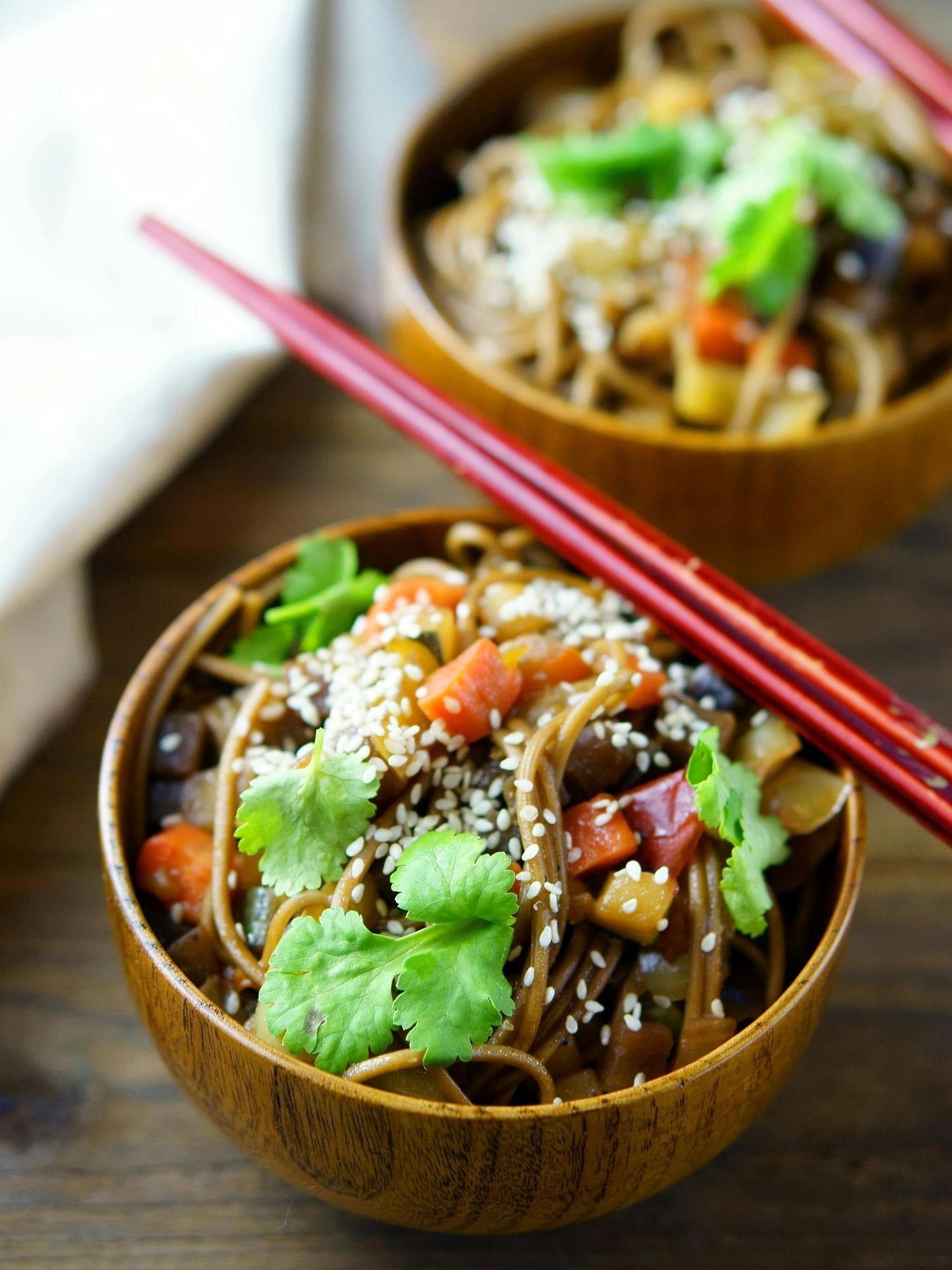 Two wooden bowls filled with stir-fried noodles and chopped vegetables, garnished with sesame seeds and cilantro. Red chopsticks rest across the foreground bowl.