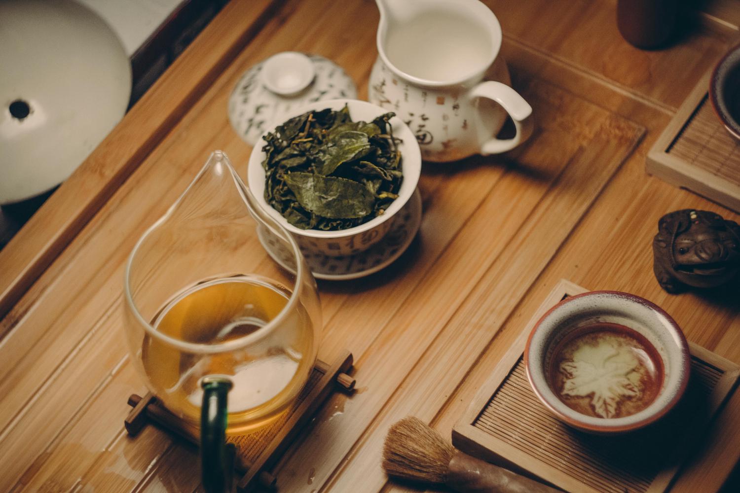 A tea set on a bamboo tray displays dried green tea leaves, a glass pot of golden brewed tea, and a teacup with a white maple leaf design. Other items include a ceramic pitcher, a small brush, and a decorative frog.
