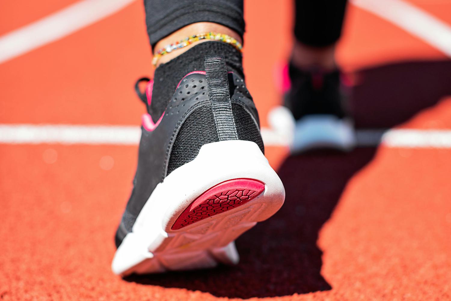 Close-up of a person's black, white, and pink running shoe on an orange track with white lines. A colorful beaded anklet is visible above the shoe.