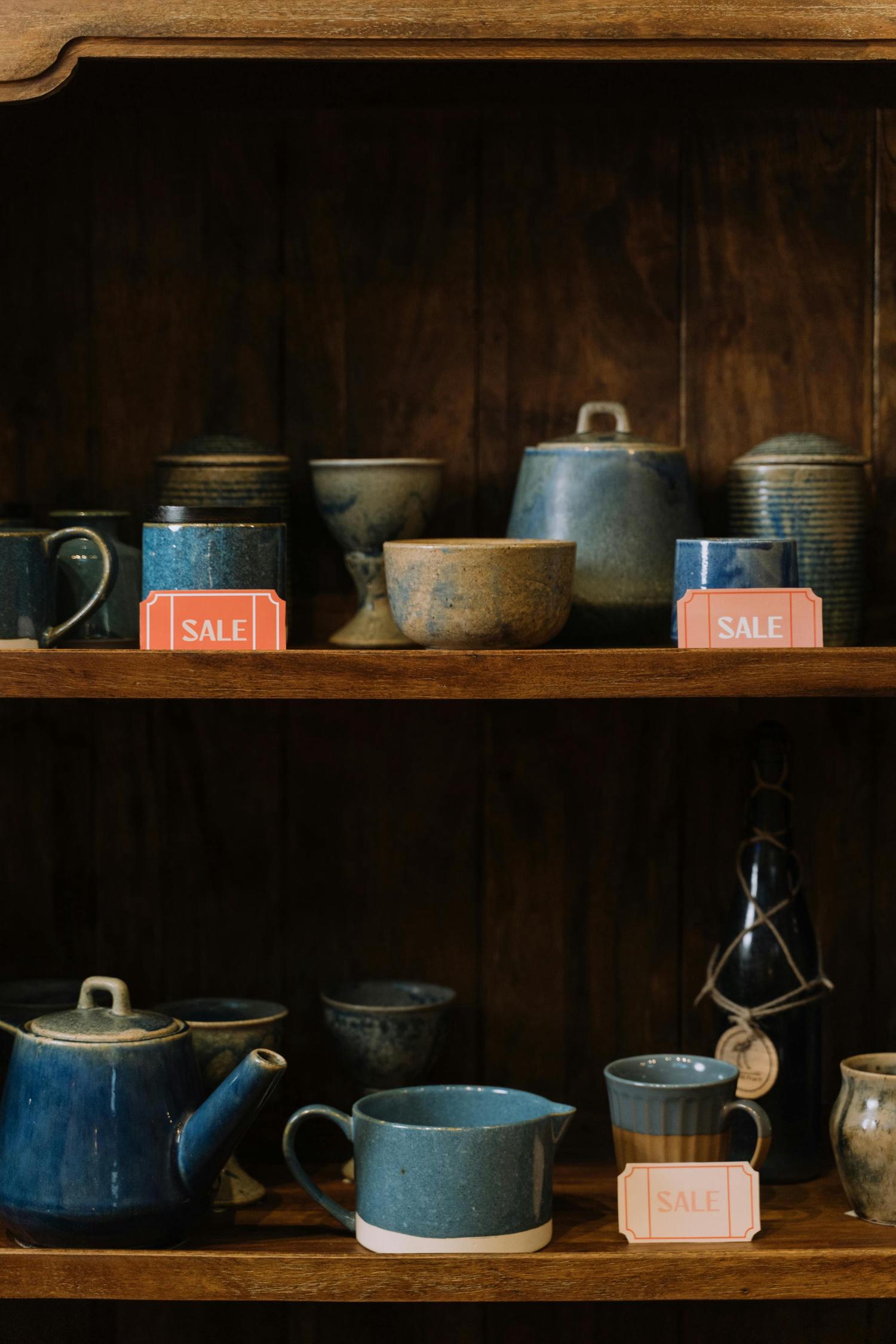 Handcrafted ceramic pottery in shades of blue and earthy tones, including mugs, bowls, and a teapot, are displayed on dark wooden shelves with several items featuring orange 