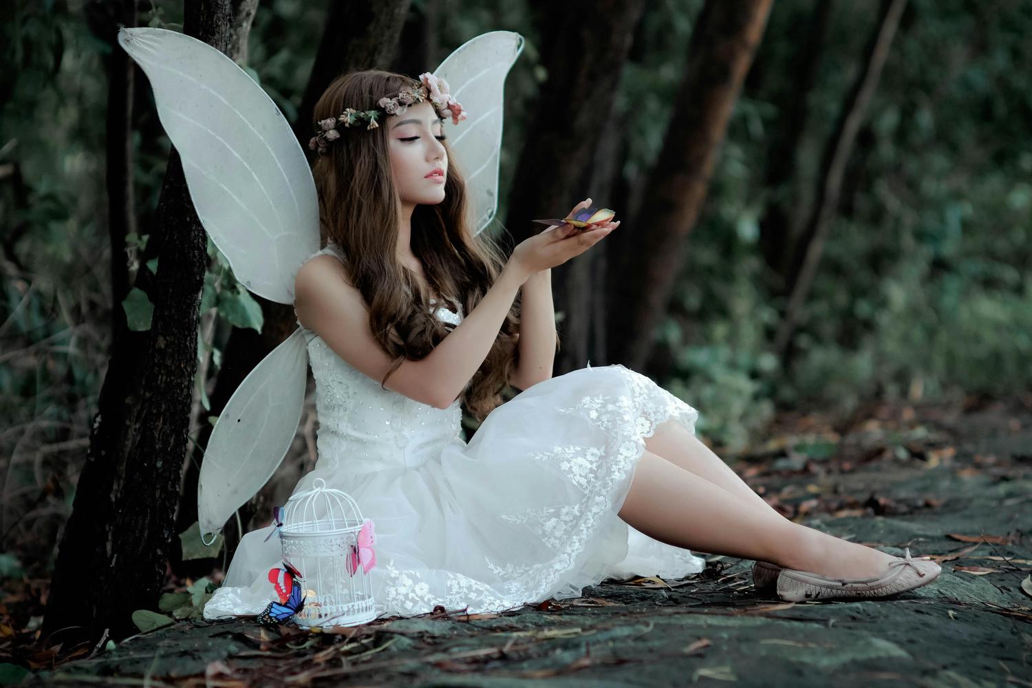 A woman dressed as a fairy, with large white wings and a floral crown, sits on the forest floor, gazing down at a small colorful butterfly or object in her cupped hands. A white decorative birdcage adorned with paper butterflies sits next to her.