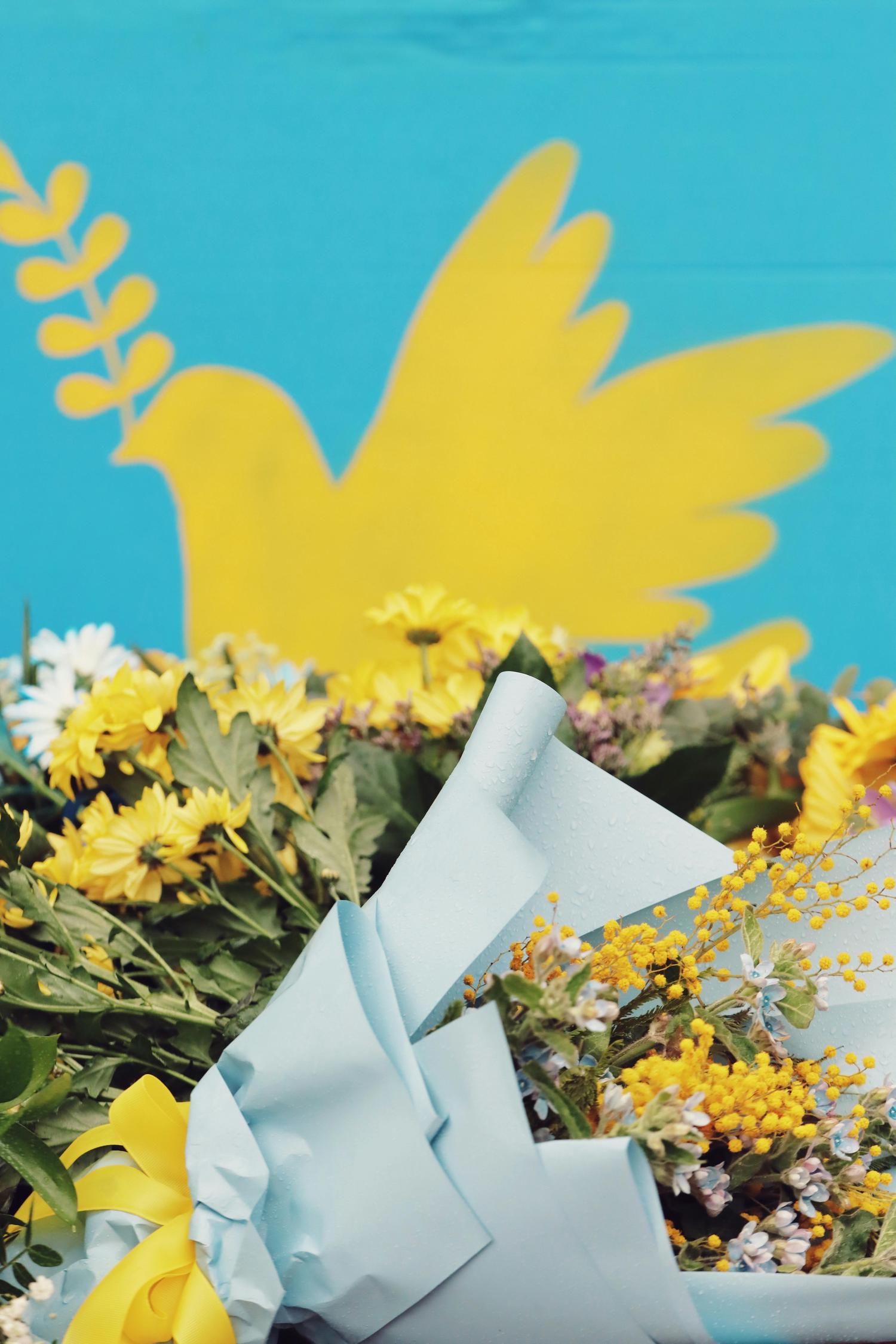 A vibrant bouquet of yellow and blue flowers, wrapped in blue paper with a yellow ribbon, rests against a blue background featuring a yellow peace dove with an olive branch. The blue and yellow colors evoke the Ukrainian flag.