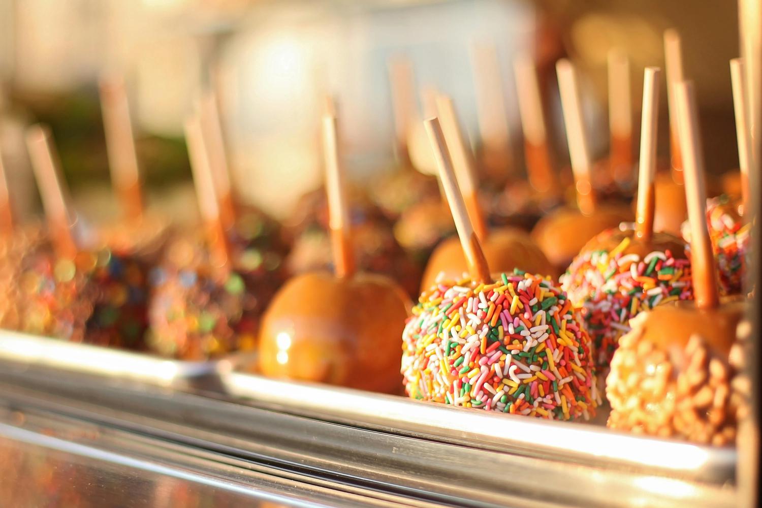 A row of caramel apples on sticks sits in a display case, with one in the foreground covered in colorful sprinkles. Other apples are visible in the blurred background, some plain and some with various toppings, all bathed in warm light.