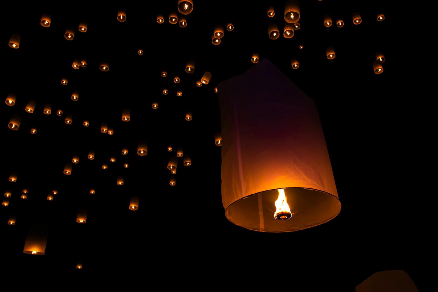 Numerous illuminated sky lanterns drift through a dark night, with one large lantern in the foreground showing its bright orange flame.