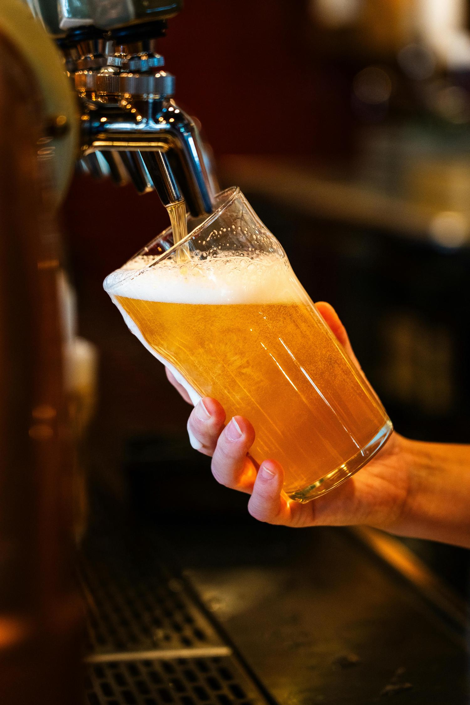 A hand holds a glass being filled with golden beer from a tap, creating a frothy white head.