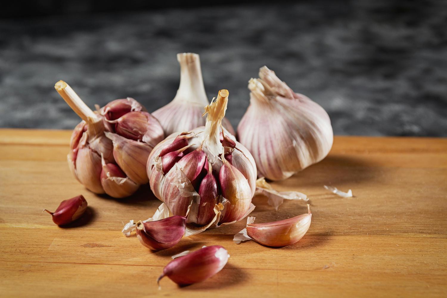 Three bulbs of garlic, some showing reddish-purple cloves, and several loose cloves are arranged on a light wooden surface against a dark background.