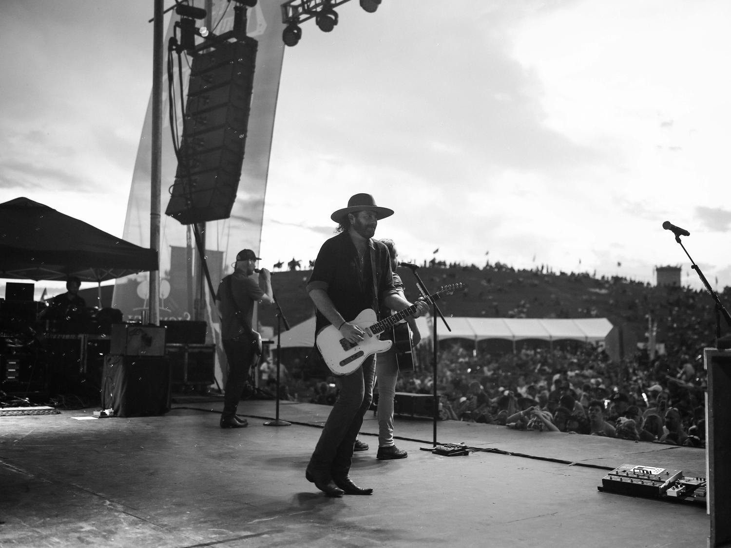 In black and white, a musician in a hat plays an electric guitar on an outdoor stage, with another musician singing and a large crowd gathered in the background under a cloudy sky.