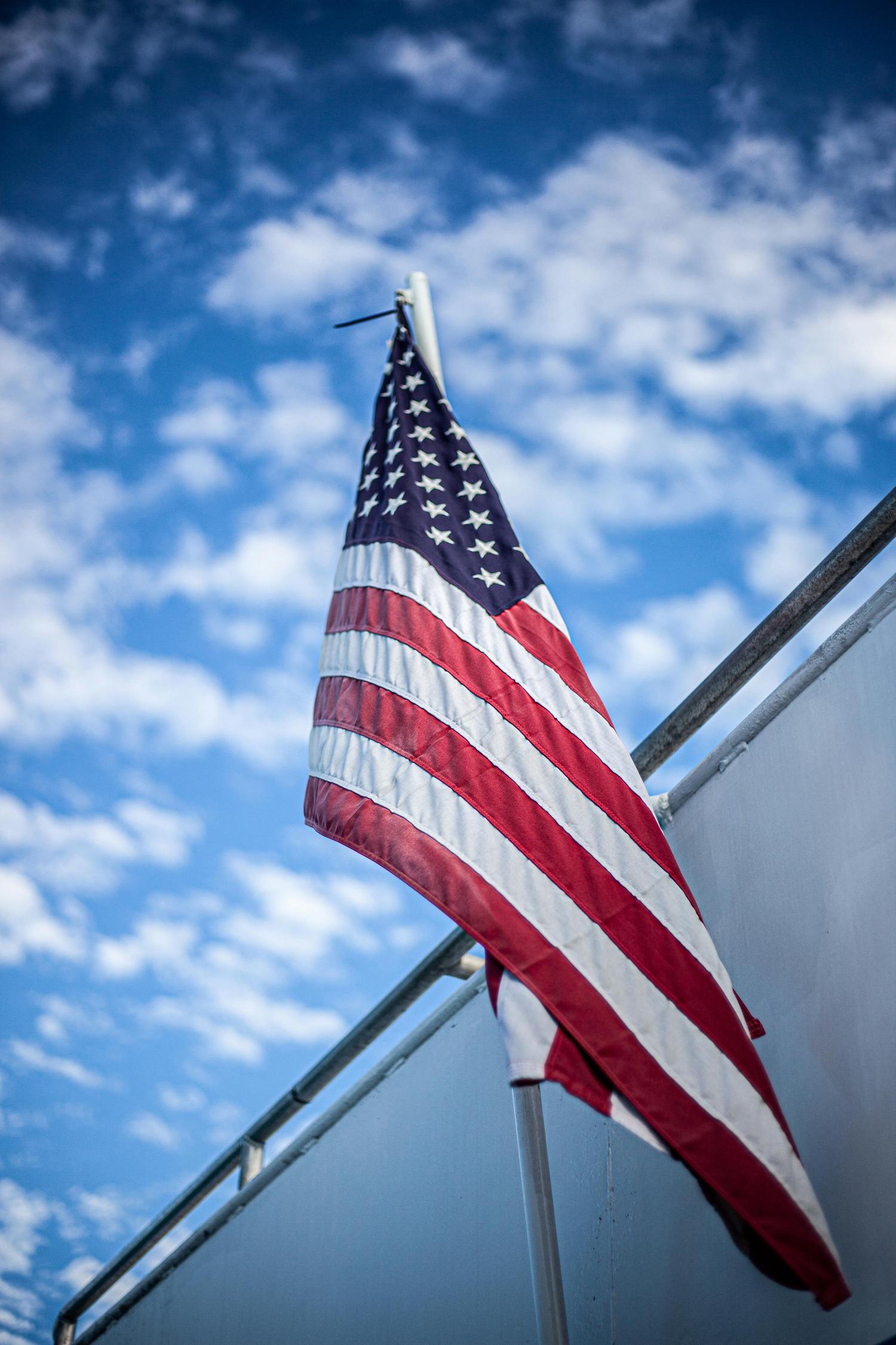 An American flag hangs from a pole, with its red and white stripes and blue field with white stars visible against a clear blue sky dotted with white clouds. A white railing partially obscures the lower part of the flag.