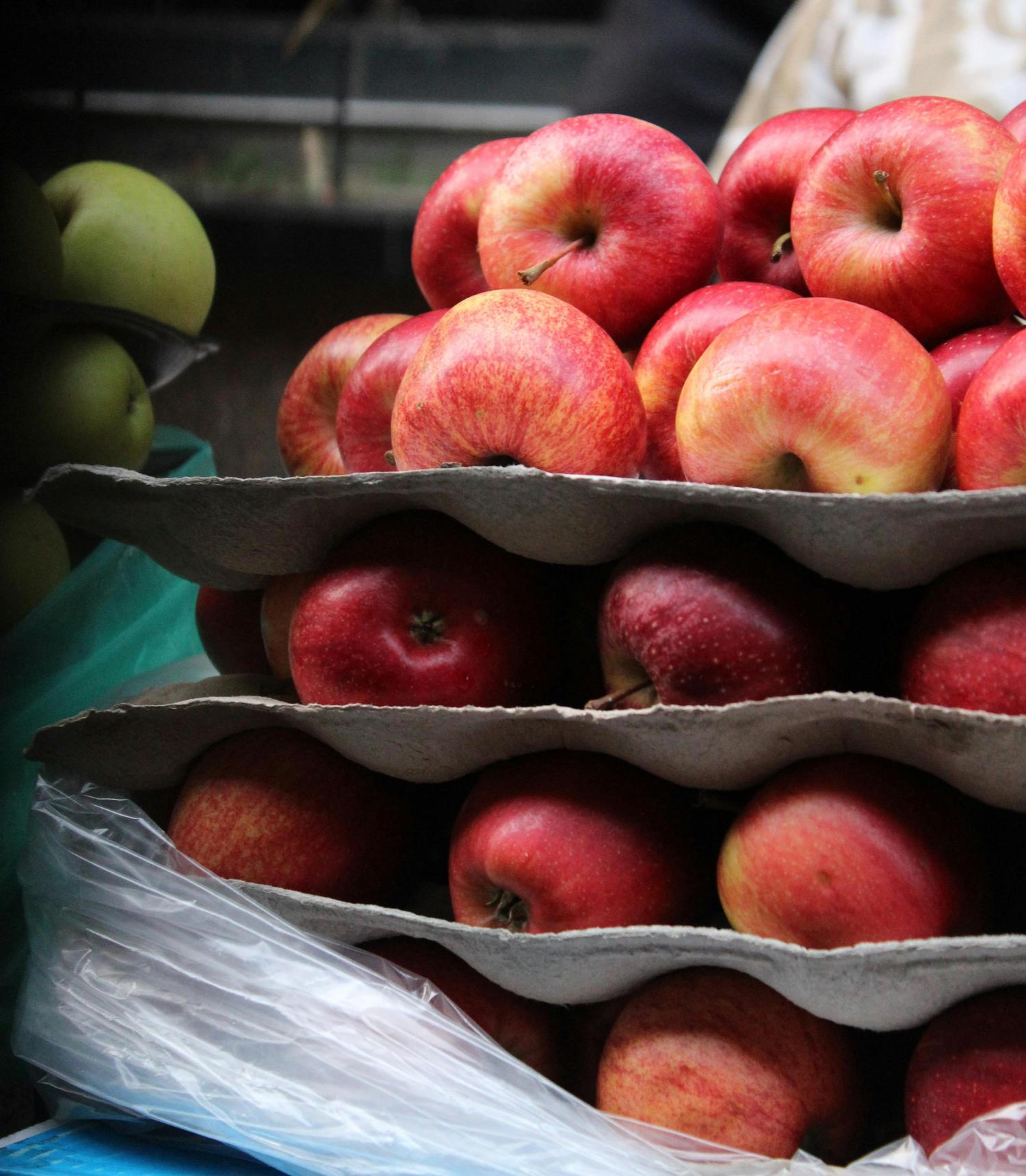 Red and yellow speckled apples are stacked in gray, molded cardboard trays. A few green apples are visible in the blurred background, along with plastic packaging.