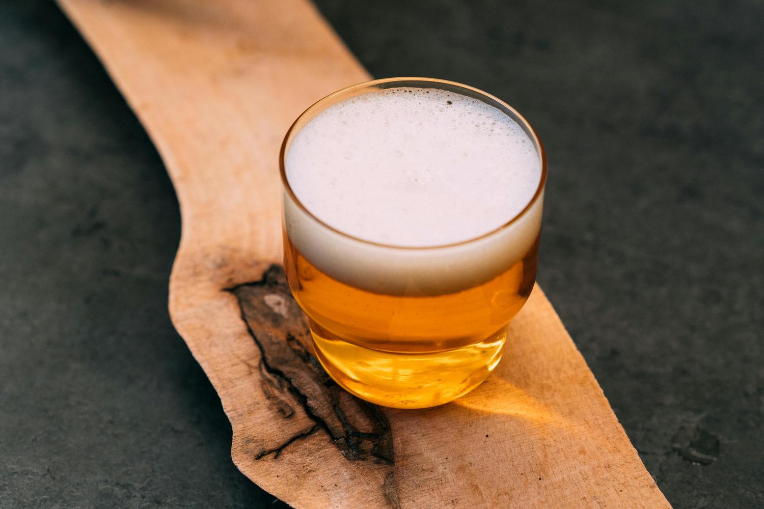 A glass of golden beer with a thick white foam head sits on a natural wooden board featuring dark grain patterns, against a dark, textured background.