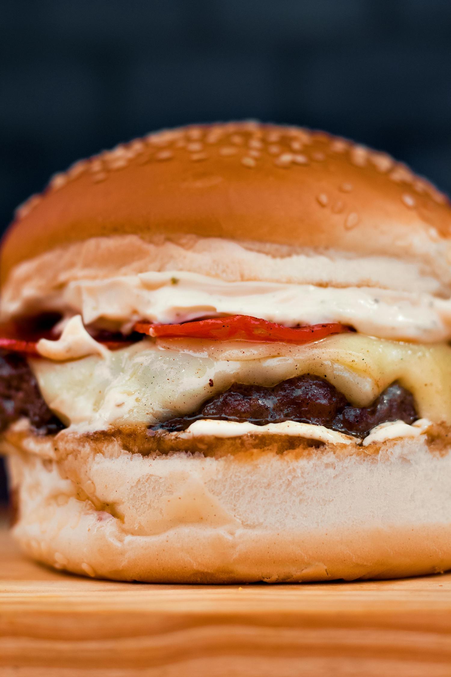 A close-up of a loaded burger featuring a sesame seed bun, melted cheese, a thick patty, red tomato slices, and creamy white sauce, sitting on a wooden surface.