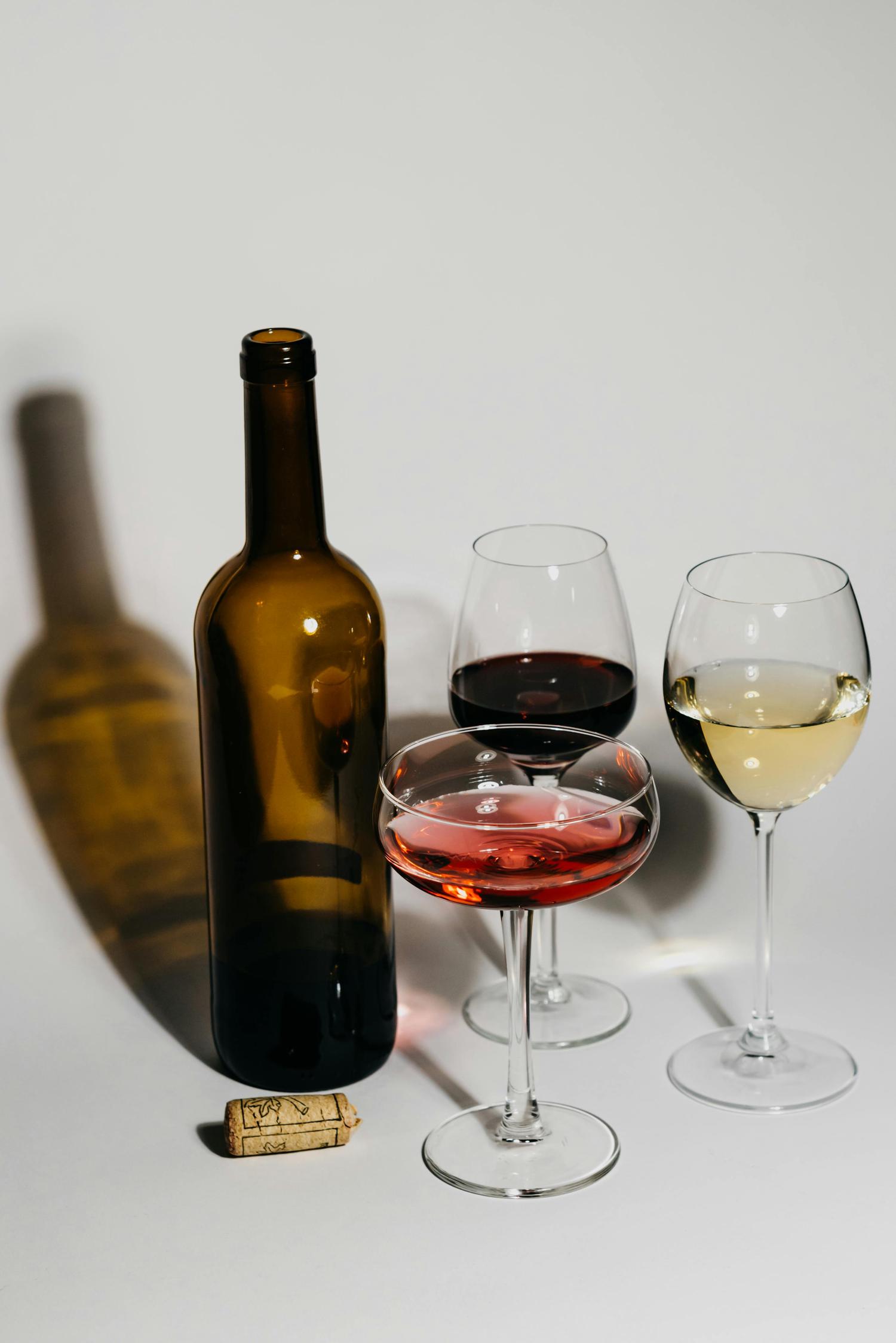 A dark brown wine bottle stands beside three wine glasses, one holding red, another rosé in a coupe, and a third with white wine. A wine cork lies on the white surface nearby.