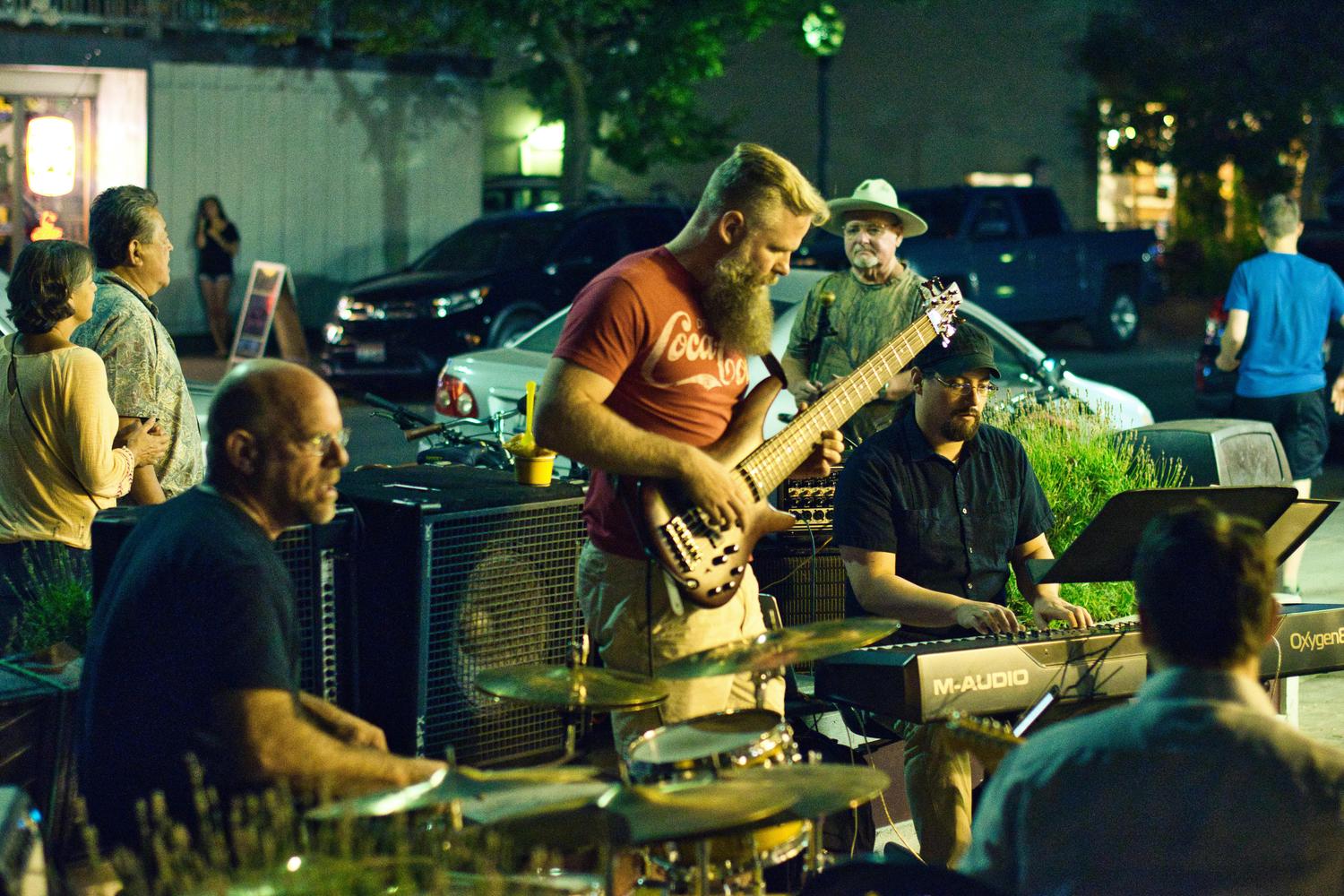 A band performs outdoors at night, with a bearded bassist in a red t-shirt playing in the foreground, alongside a keyboardist and drummer. Other musicians and a few onlookers are visible in the background under streetlights.