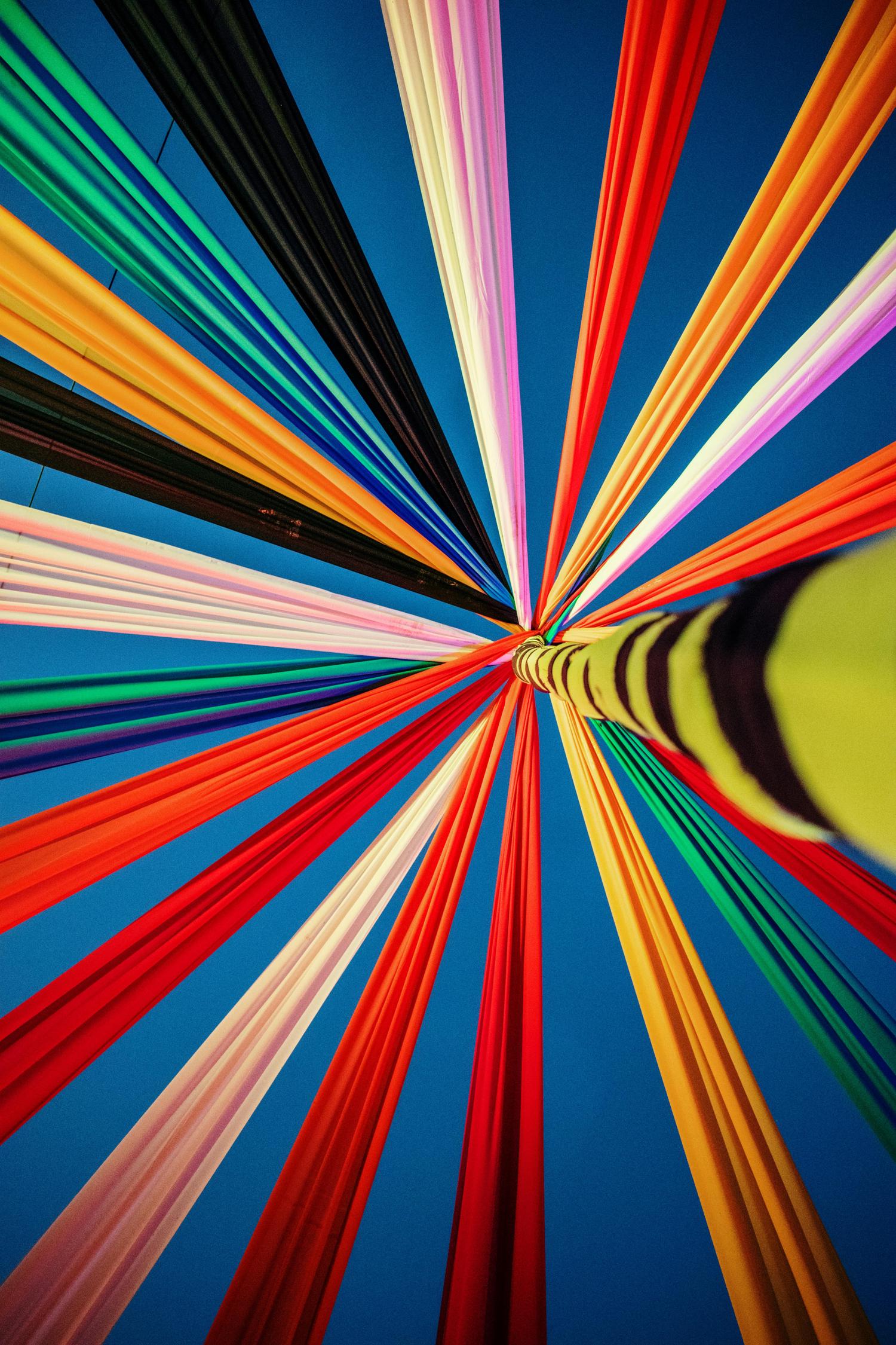 Colorful fabric strips in various shades radiate outwards from a central point, forming a vibrant canopy against a clear blue sky. The fabrics include red, orange, yellow, green, blue, black, pink, and white, with the central structure wrapped in green and black stripes.