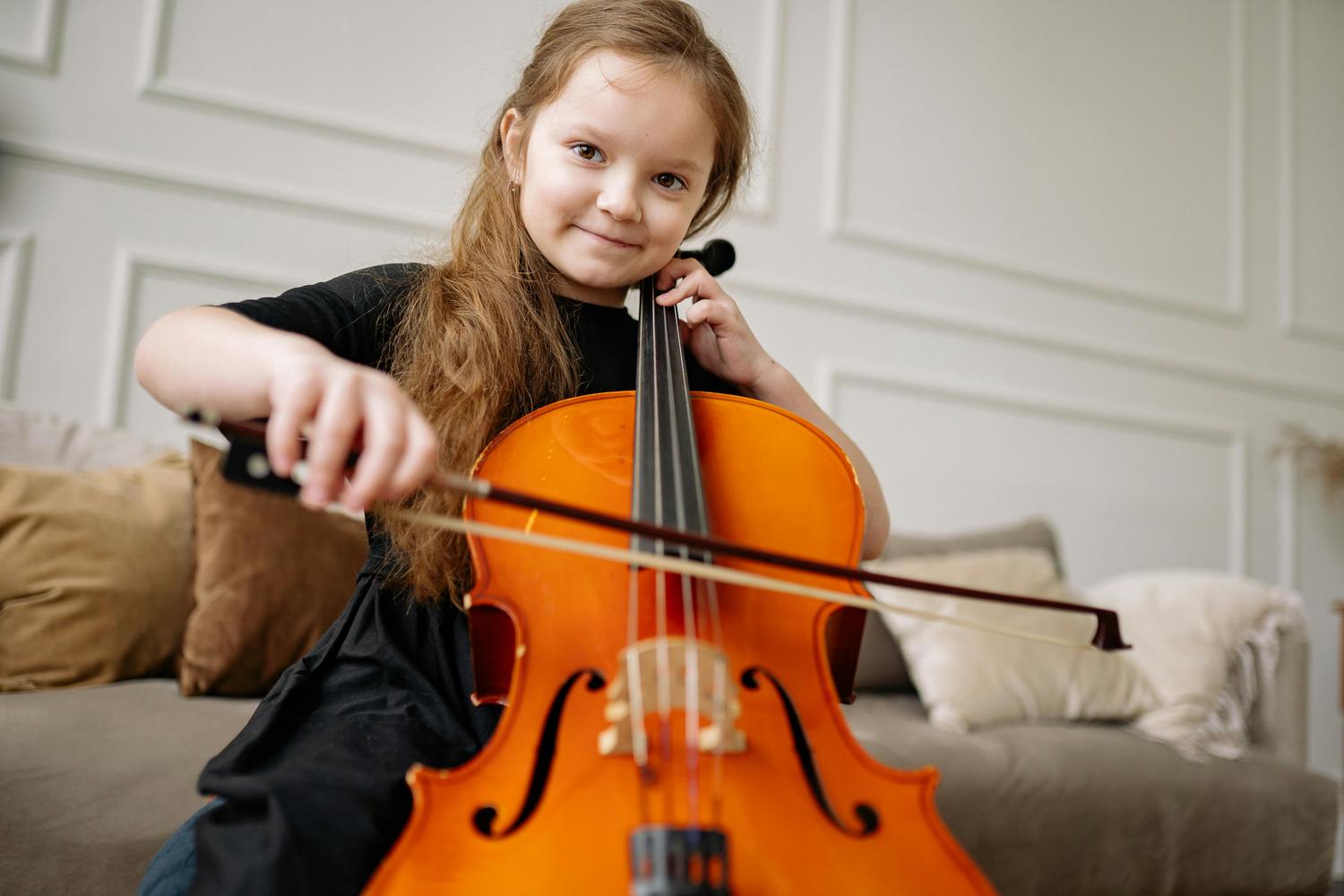 A young girl with long brown hair smiles at the camera while holding a cello and its bow.