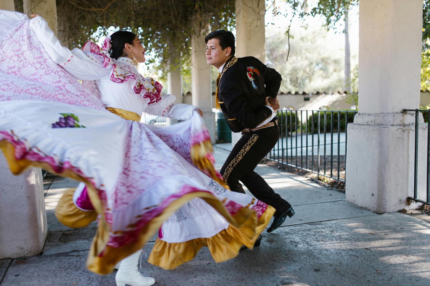 A female and male dancer perform outdoors in traditional attire; her white skirt with pink patterns billows widely in motion, while he strikes a dynamic pose in a black charro suit with gold embroidery.