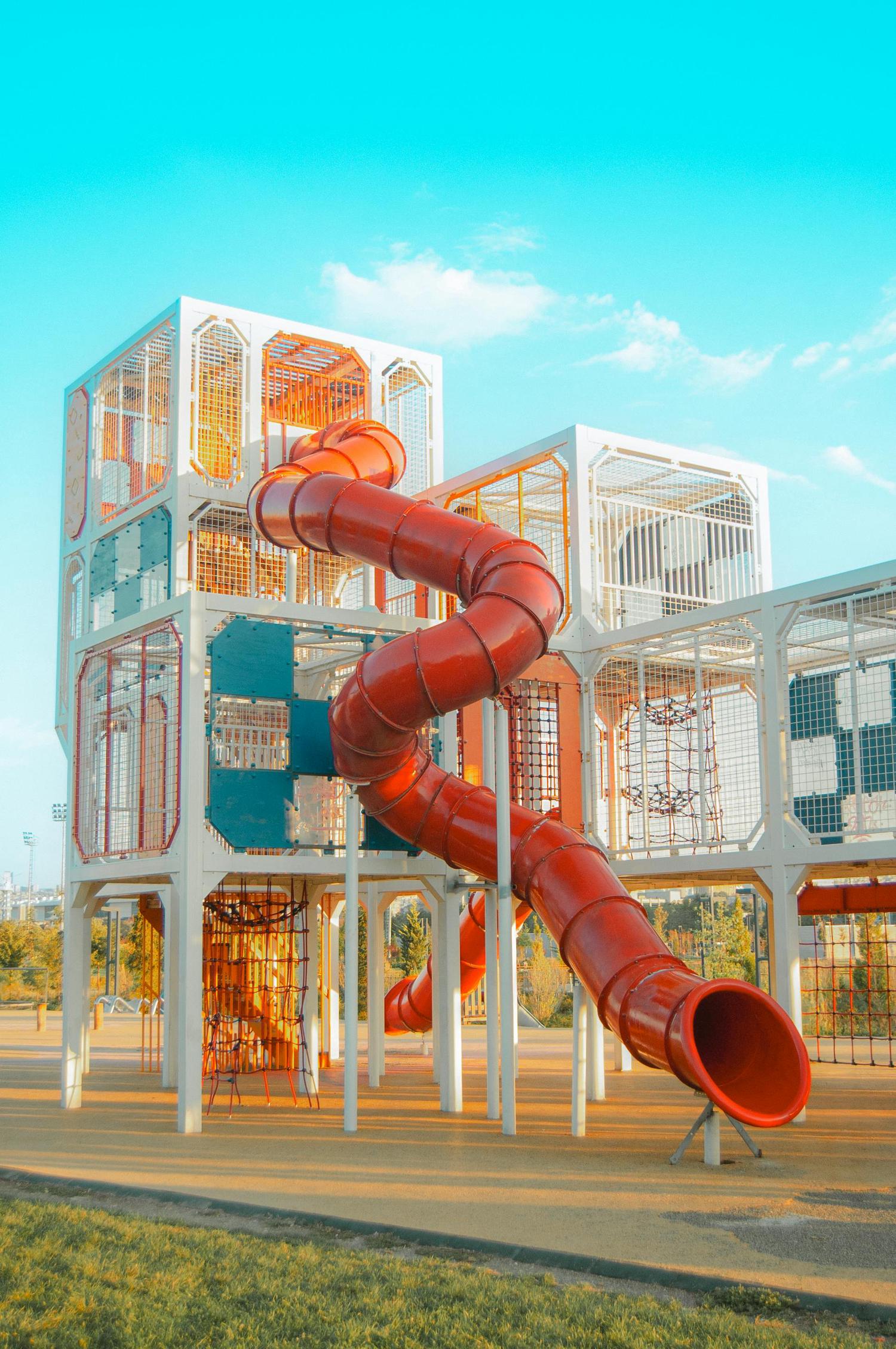 A modern, multi-level playground structure with white frames, orange and teal accents, and a winding red tube slide. It sits on a light-colored ground surface with green grass in the foreground, under a bright blue sky.