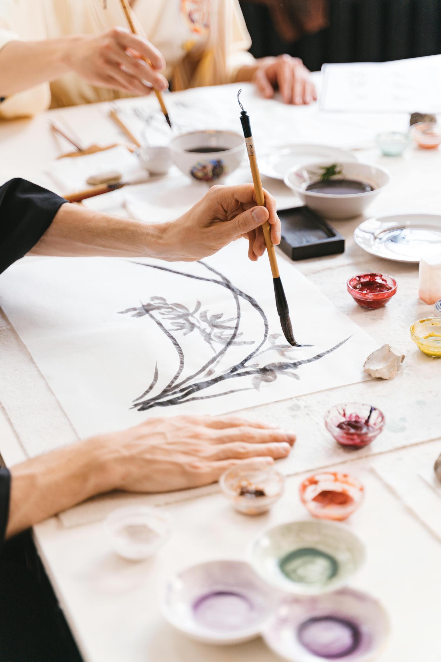 A person's hand uses a calligraphy brush to paint black ink branches and leaves on white paper. Several small bowls of colorful pigments and more brushes are scattered across the white tabletop.