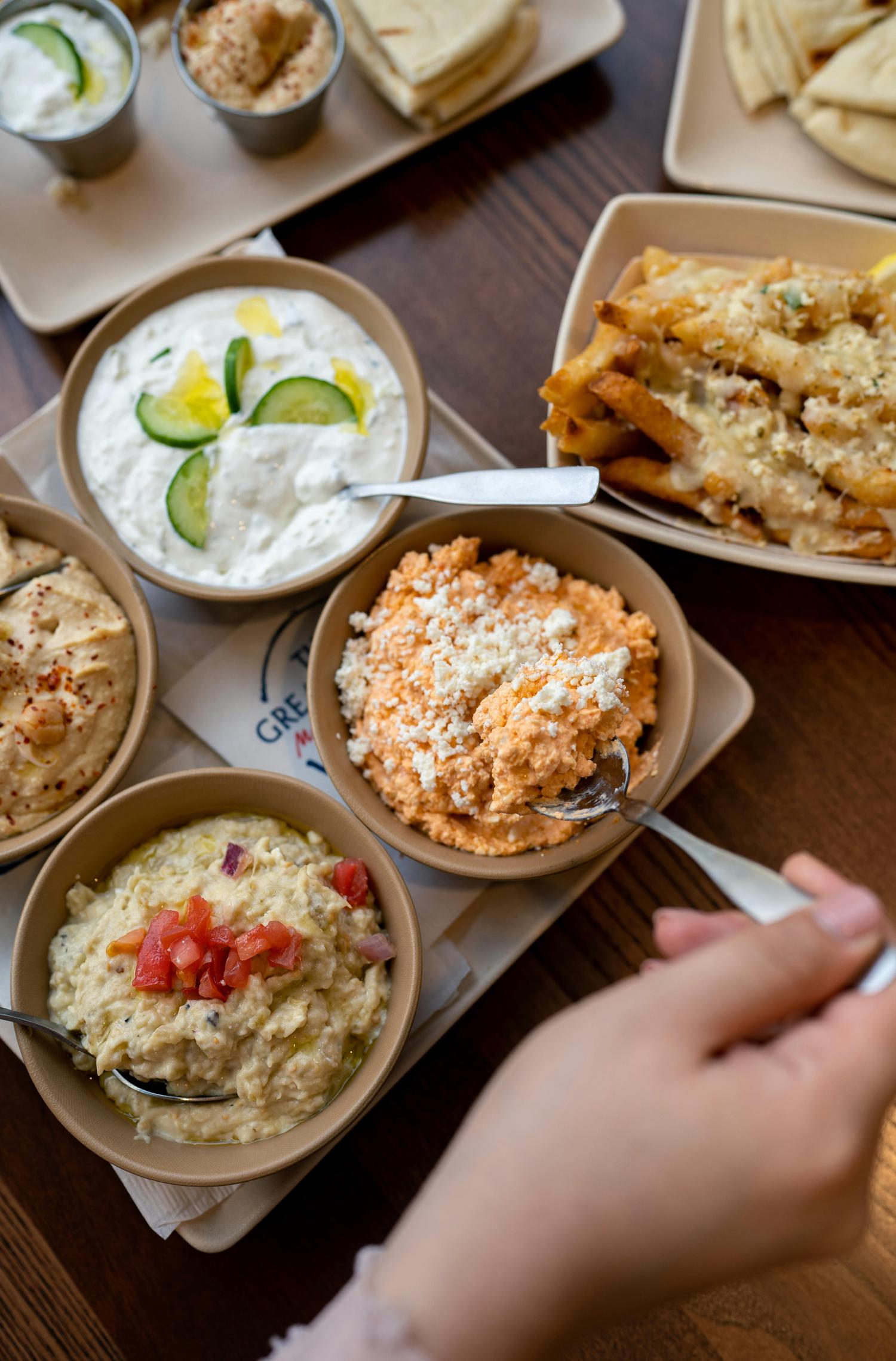 A wooden table displays an assortment of Greek dishes, including bowls of tzatziki, hummus, spicy feta dip, and baba ghanoush, along with cheesy fries and pita bread. A hand uses a spoon to scoop a portion of the spicy feta dip.