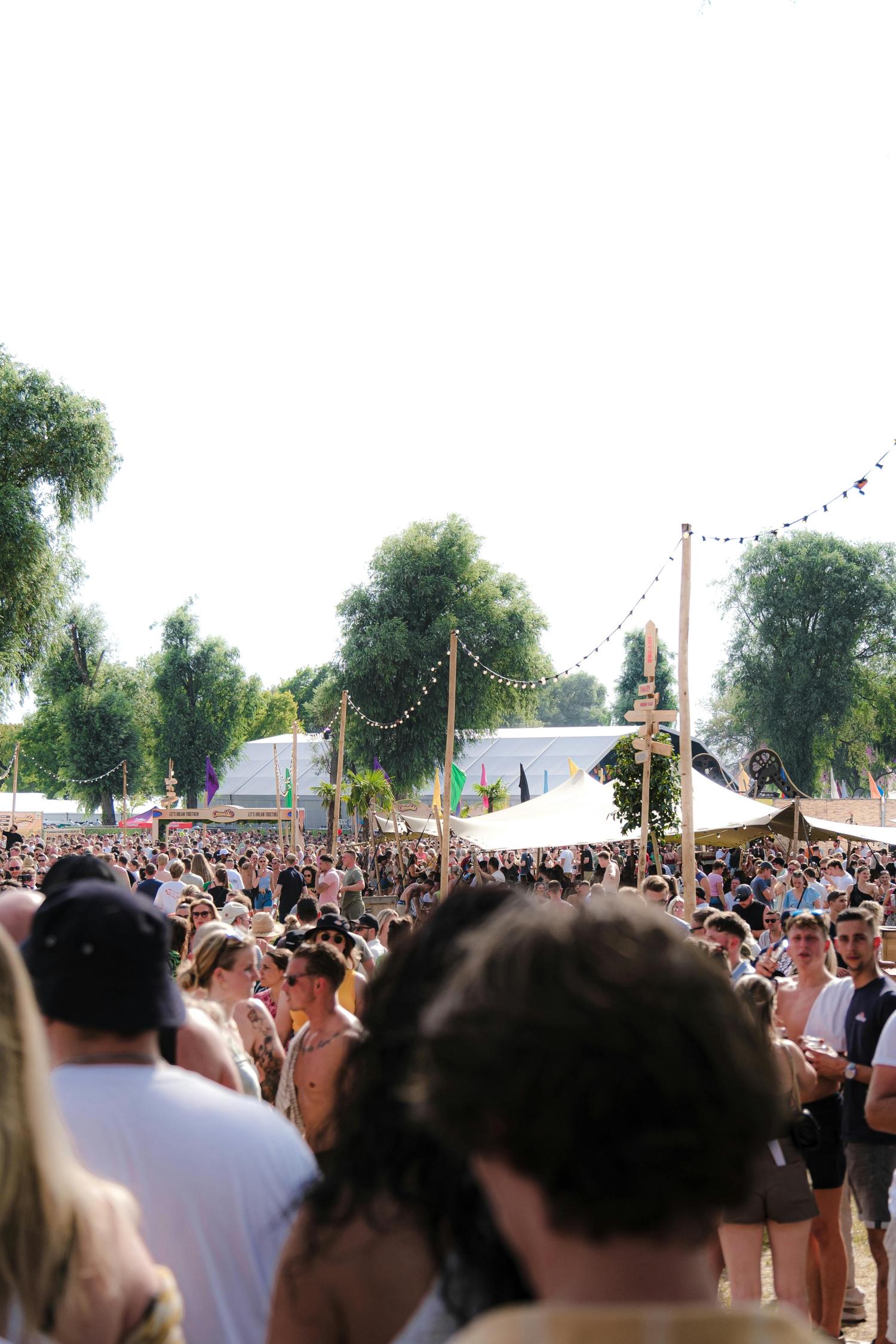 A large crowd of people gathers at an outdoor festival or event, with white tents, colorful flags, and string lights visible amongst the trees. A wooden signpost offers directions to various attractions.