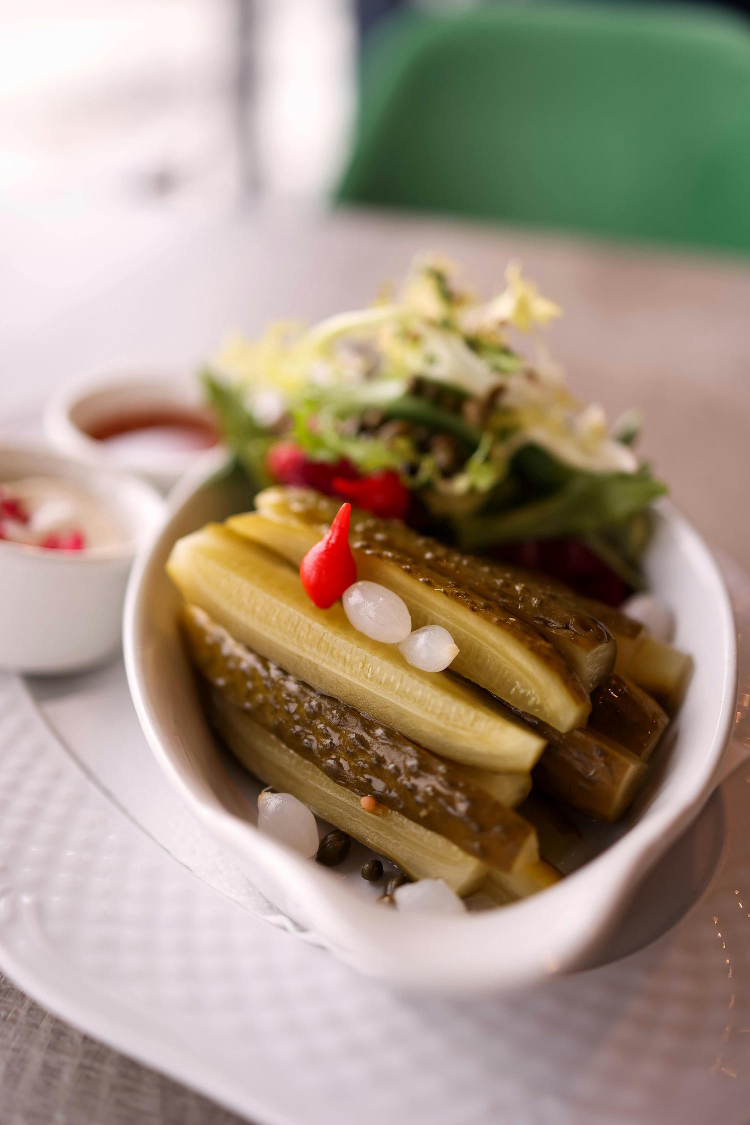 A white bowl of sliced pickled cucumbers, garnished with small white onions and a red chili pepper. It is served with a green side salad and two dipping sauces on a textured white plate.