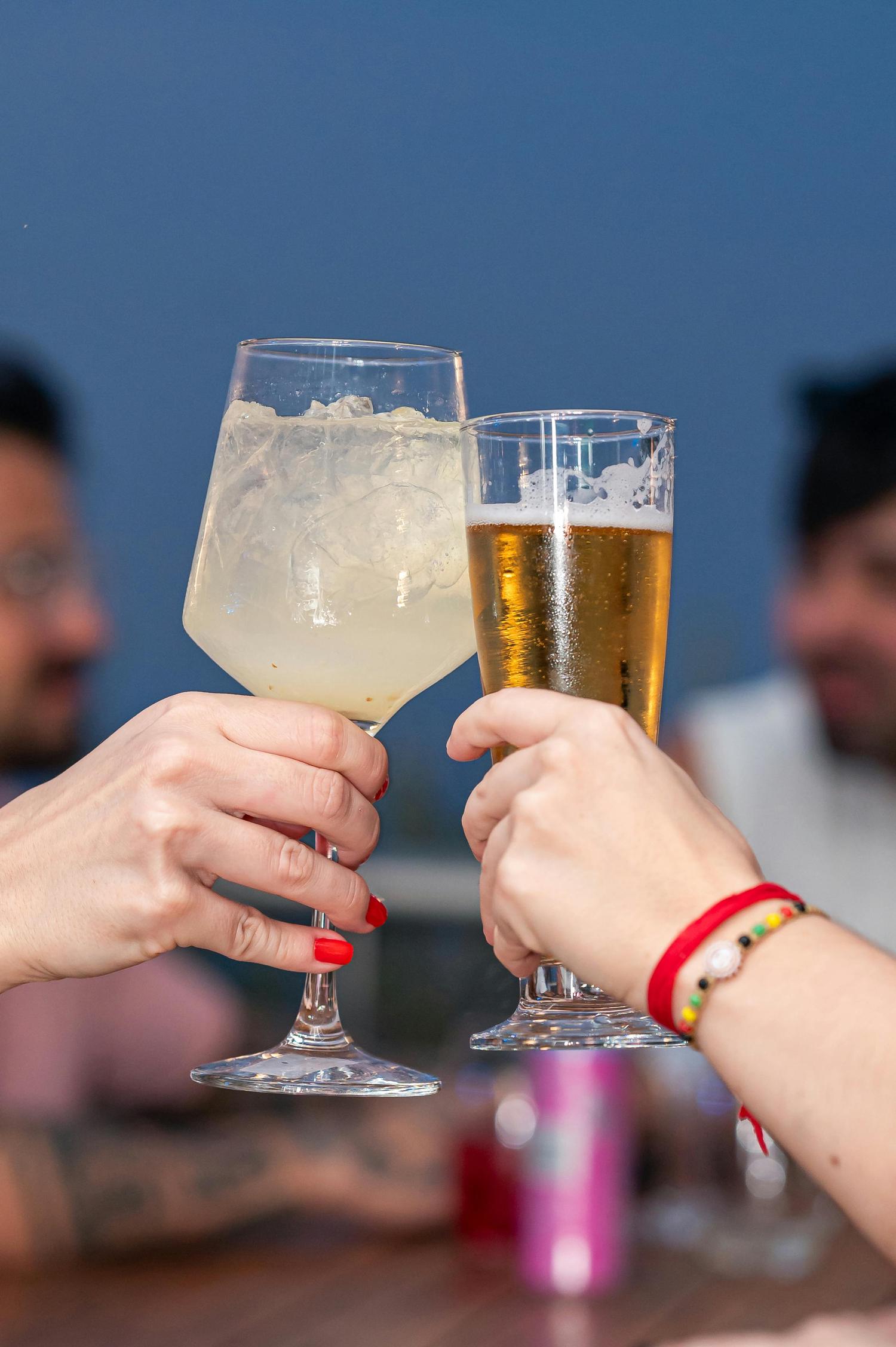 Two hands clink glasses in a toast, one holding a tall cocktail with ice and the other a glass of golden beer. The hand with the cocktail has red nail polish, while the other wears colorful beaded bracelets.