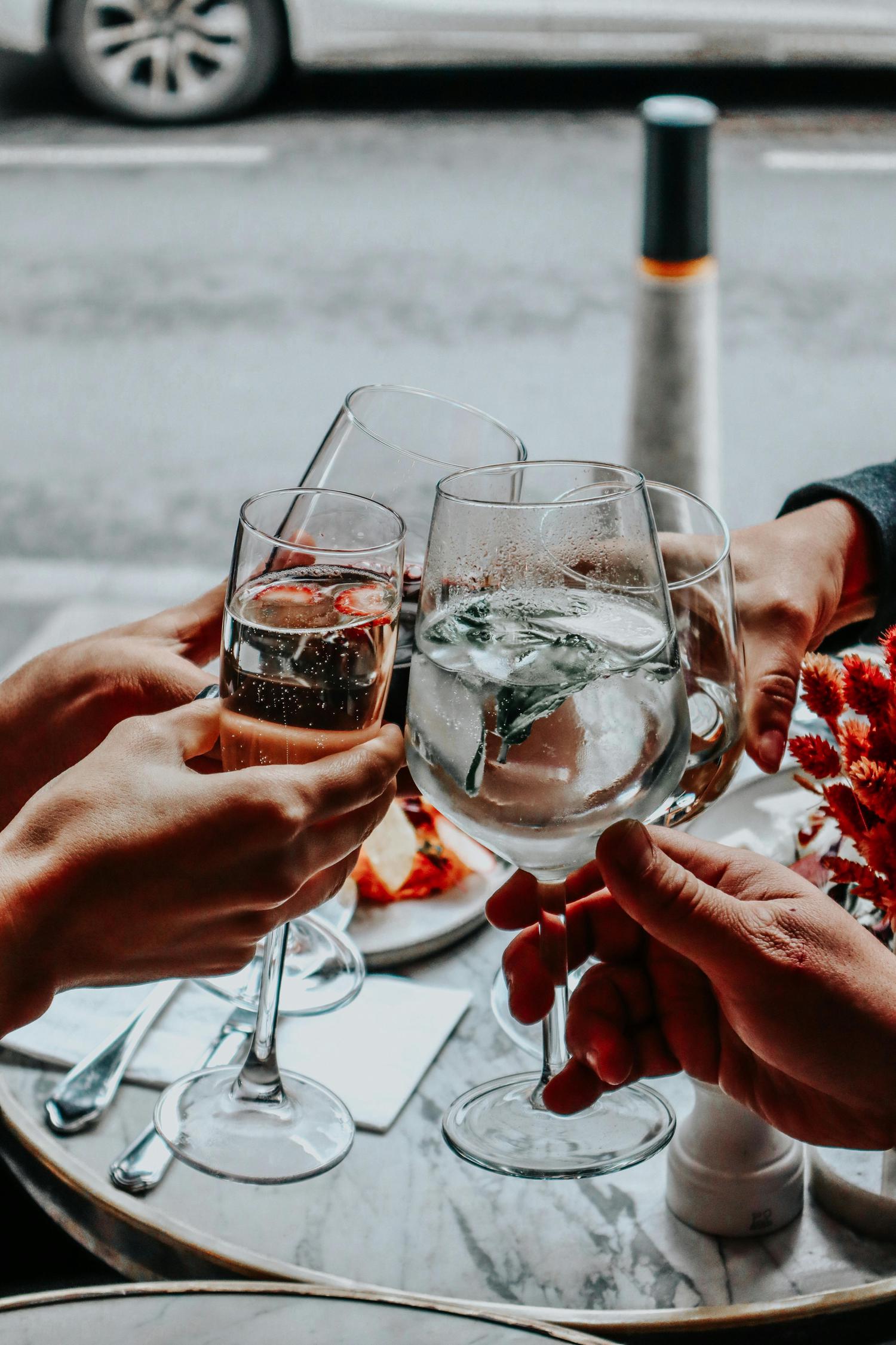 Four hands clink glasses of sparkling wine and a clear drink, toasting at a marble outdoor cafe table with a blurred street and car visible in the background.