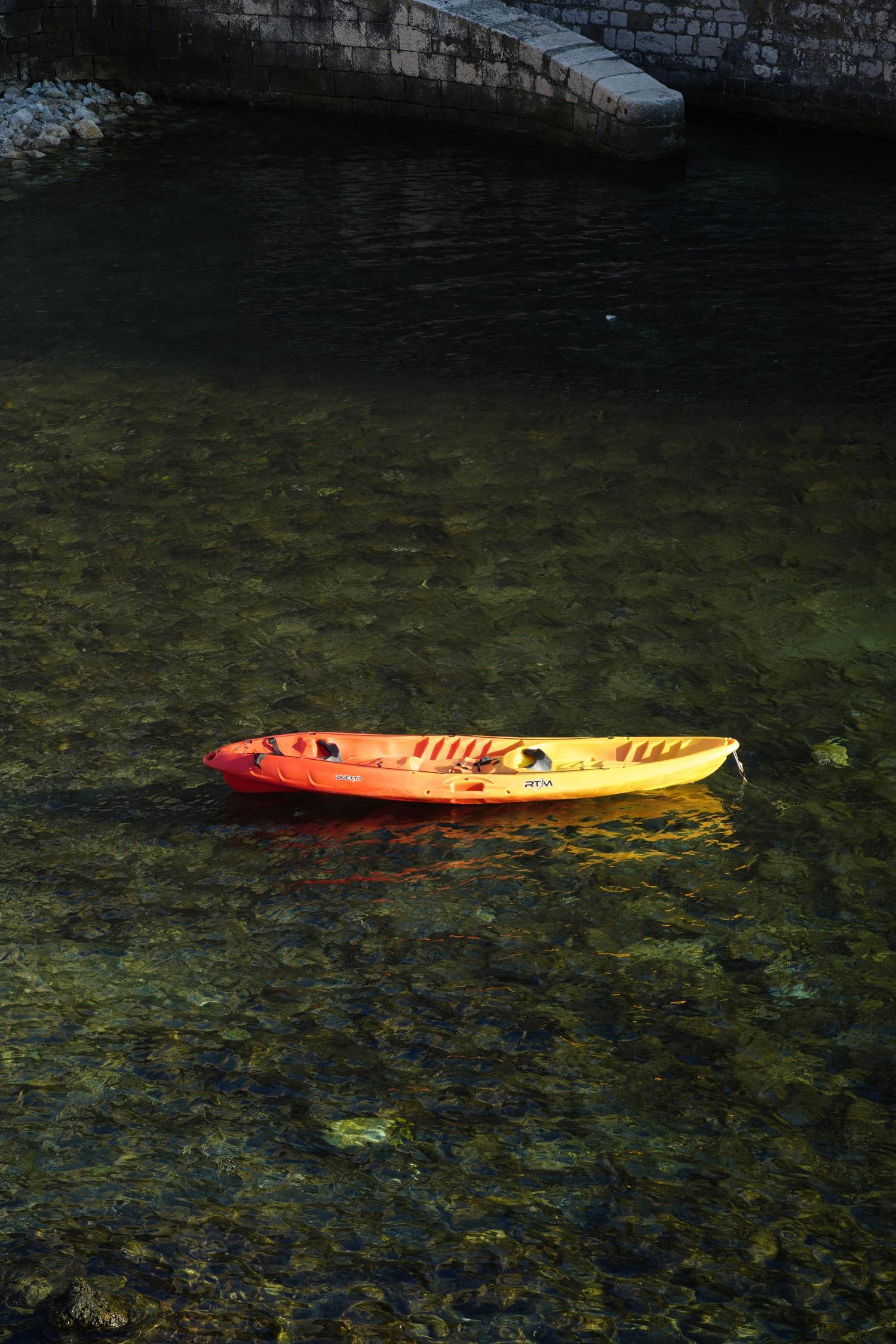 A vibrant orange and yellow kayak floats on clear, sunlit dark green water, with a stone wall visible along the far edge.