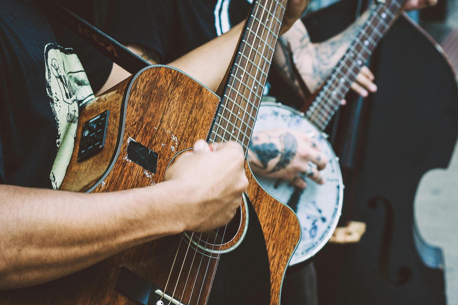 A person plays a wooden acoustic guitar with an integrated pickup, while another person's tattooed hand frets a banjo in the blurred background.