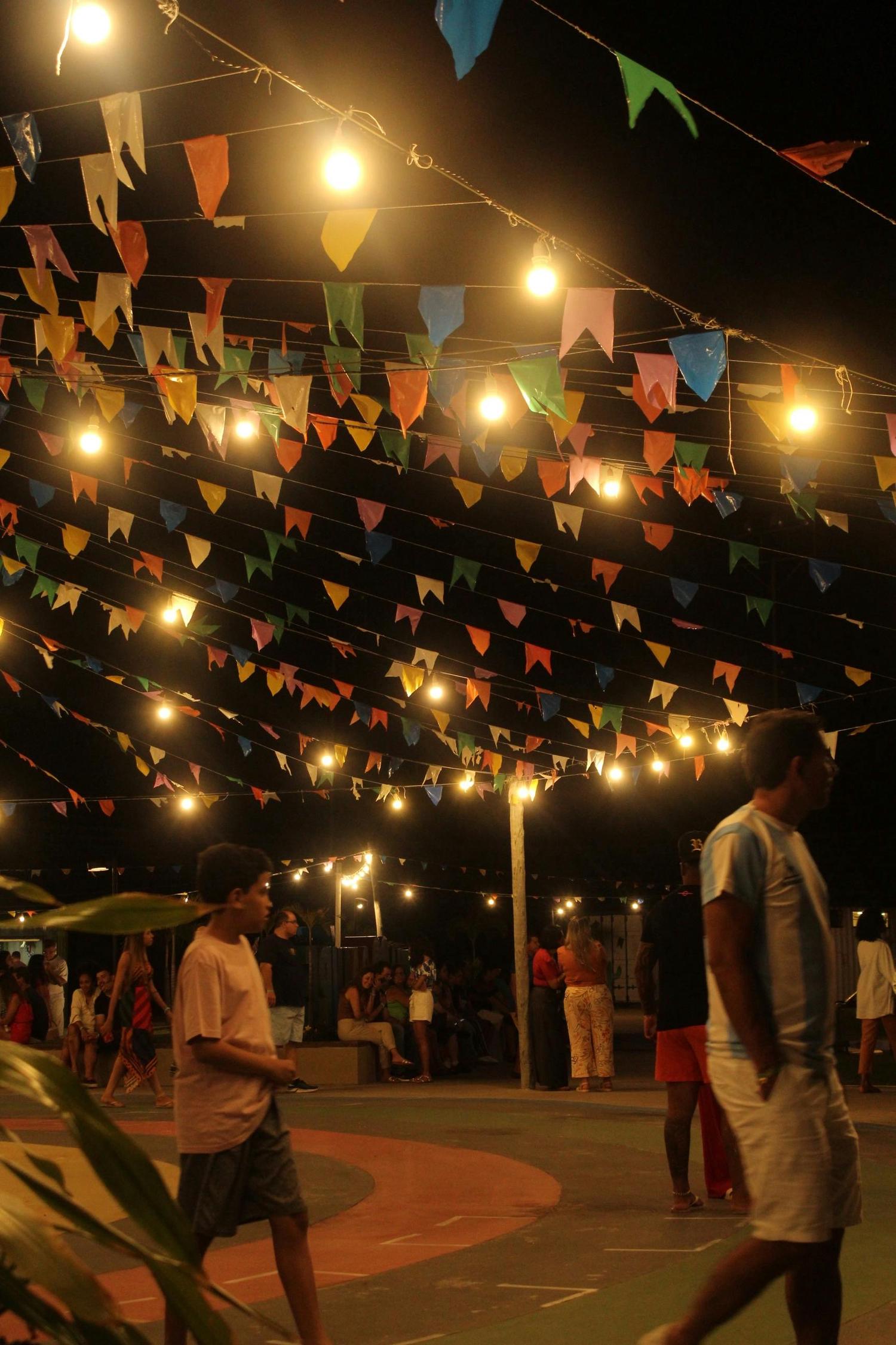 Colorful triangular flags and string lights hang overhead against a dark night sky, illuminating a casual evening gathering of people below.