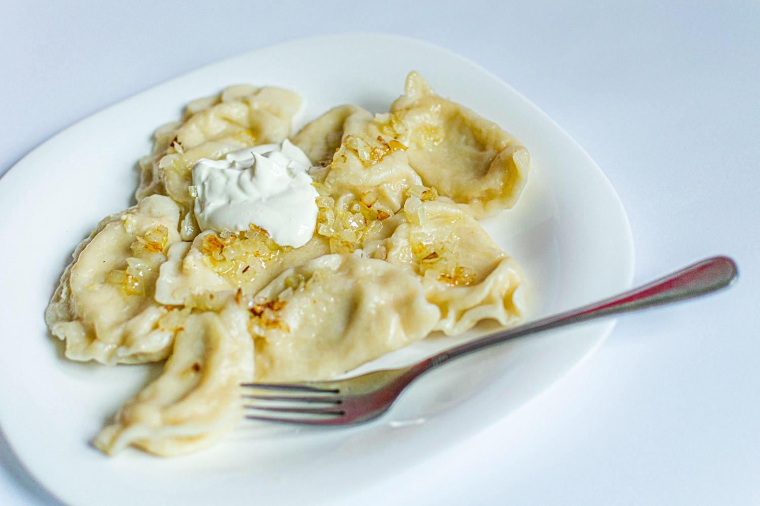 A white plate holds several golden pierogi topped with caramelized onions and a dollop of white sour cream. A silver fork rests on the right side of the plate.