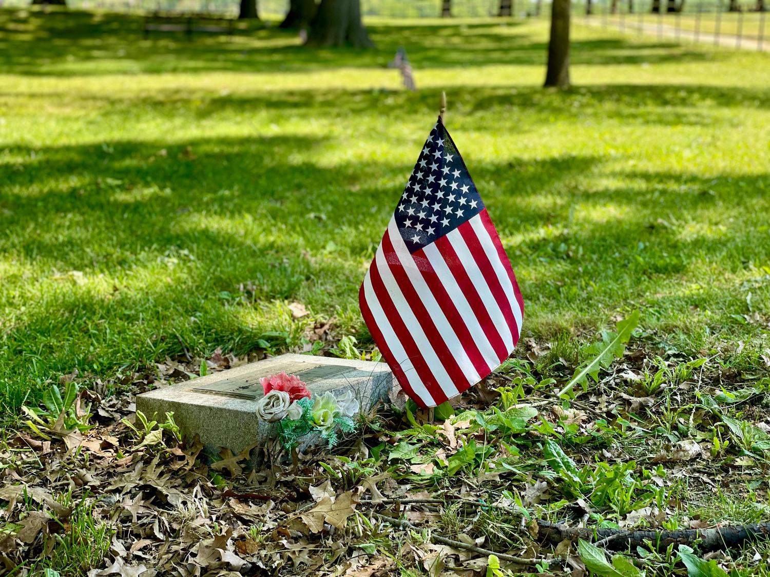 A small American flag stands next to a flat grave marker adorned with artificial flowers, set on sun-dappled grass and fallen leaves.