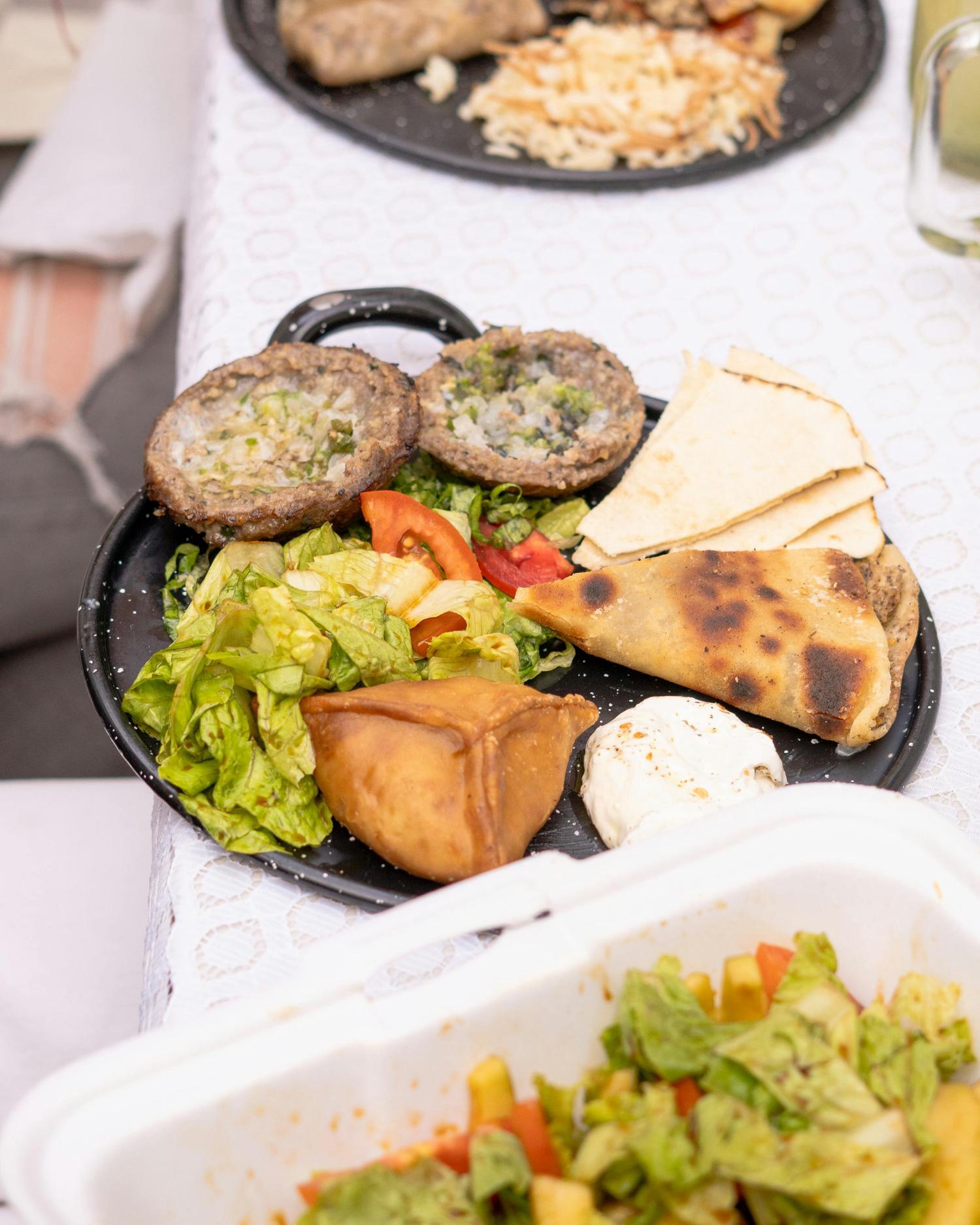A black plate holds a savory meal featuring two open-faced baked pastries with green and white filling, a fresh green salad with tomato wedges, pieces of pita bread, a golden fried pastry, a larger baked pastry, and a dollop of white dip, all on a patterned white tablecloth.