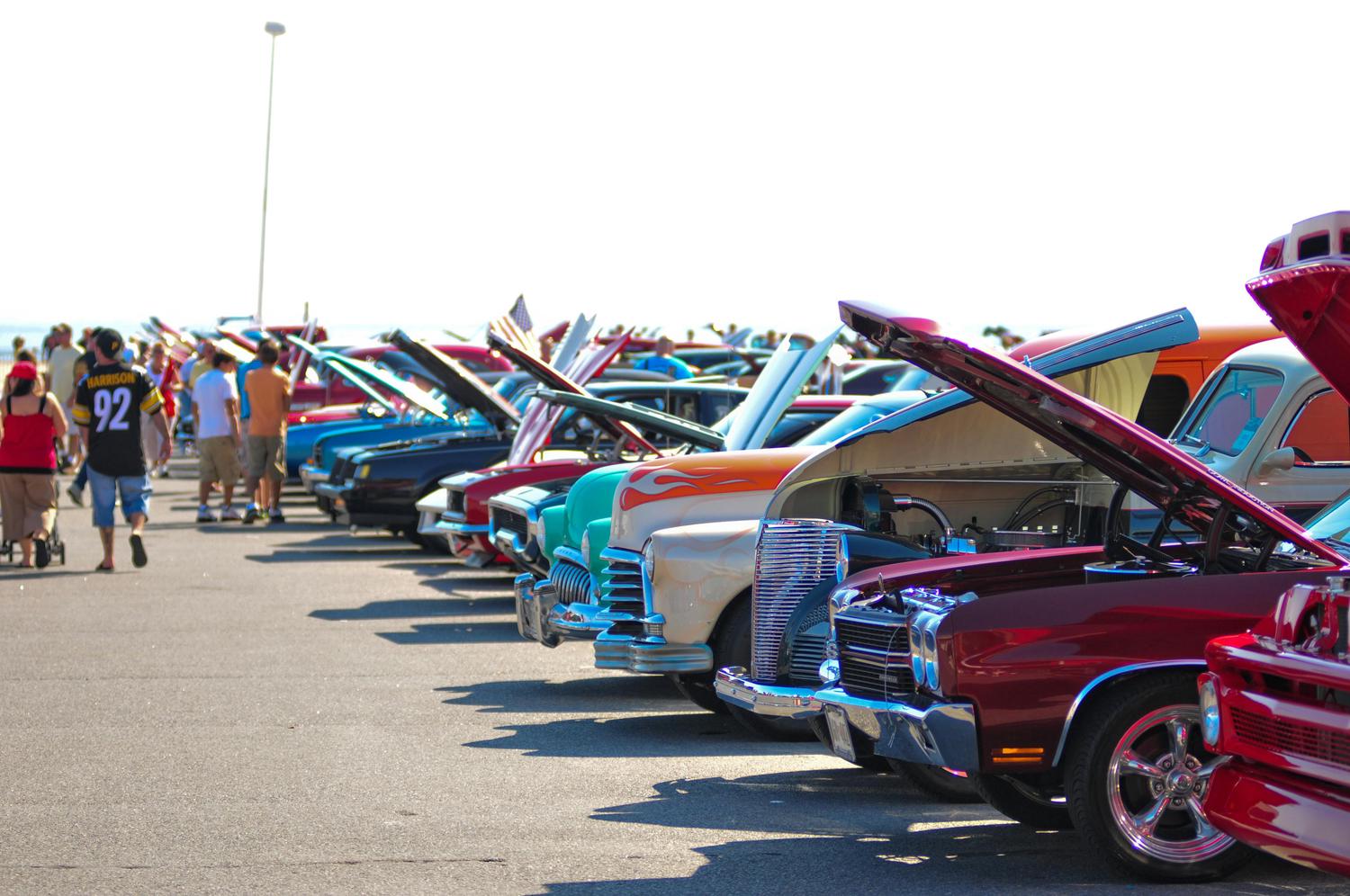 A vibrant outdoor car show features a line of classic cars with their hoods open, showcasing detailed engines and chrome grilles. Spectators stroll along the sunny asphalt lot in the background.