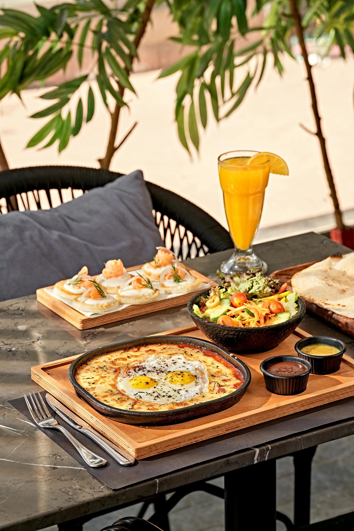 A hearty meal is presented on a dark marble table, featuring a skillet of baked eggs with melted cheese, a fresh salad, and salmon blini. Accompanying the dishes are flatbread, dipping sauces, and a glass of orange juice, set against a backdrop of green leaves.