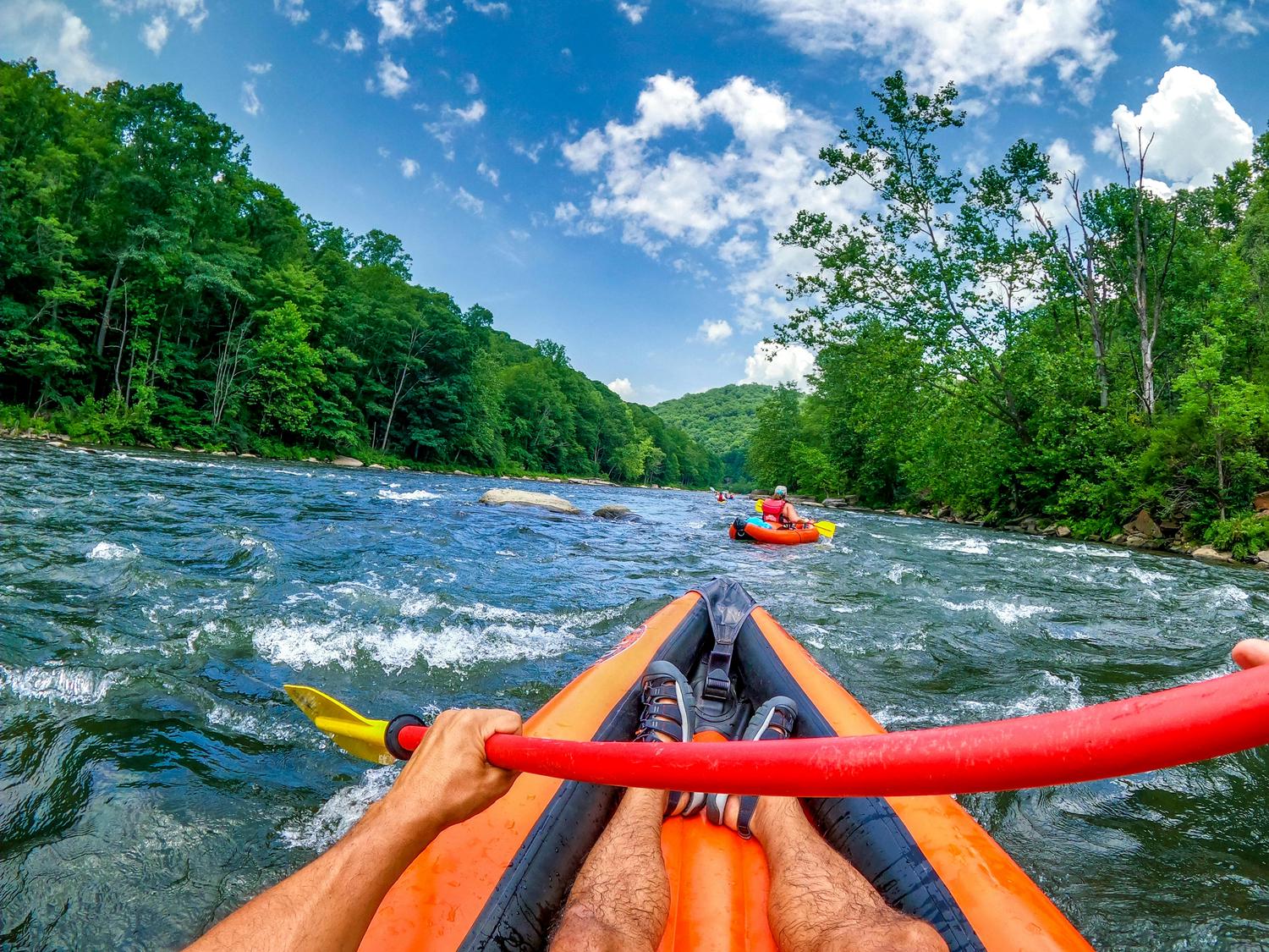 First-person view from an orange kayak on a river with rapids, a person's hands gripping a red paddle. Lush green forested mountains line the river under a blue sky with white clouds, with other kayakers visible ahead.
