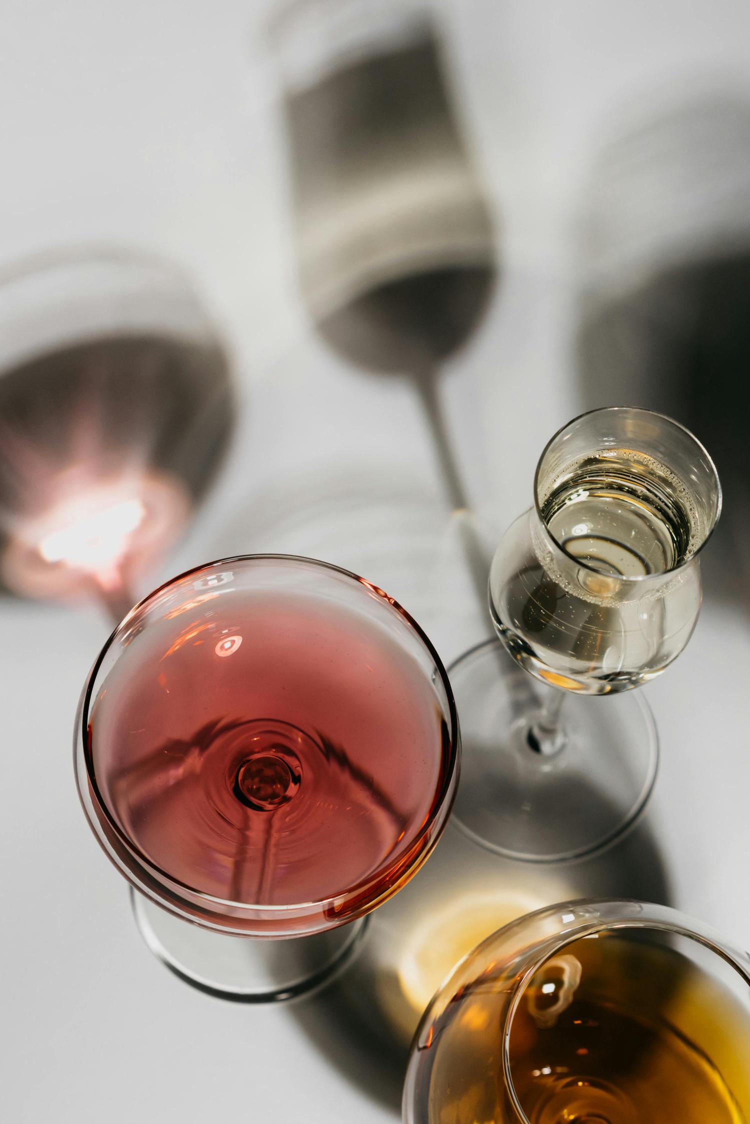 An overhead view shows three stemmed glasses filled with a reddish-pink, a sparkling clear, and an amber liquid, casting distinct shadows on a light surface.