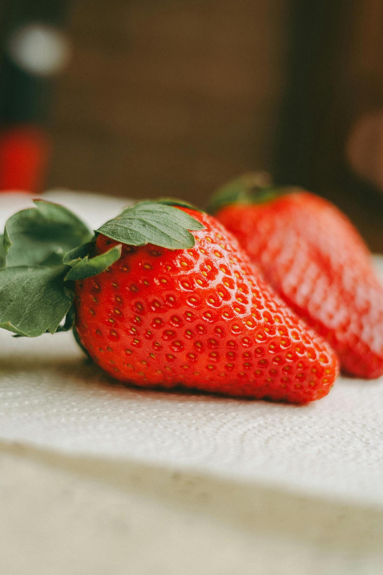 A bright red strawberry with green leaves and visible seeds rests on a textured white surface, with a second strawberry blurred in the background.