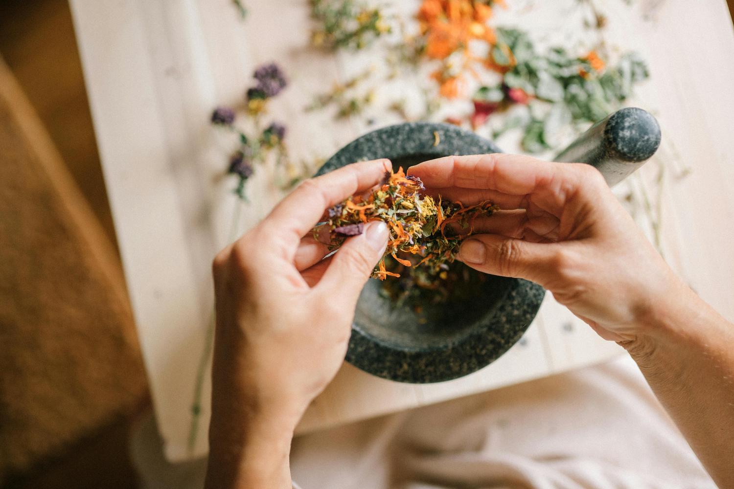 Overhead view of hands holding a mix of dried orange, green, and purple herbs and petals over a mortar, with a pestle nearby and more herbs scattered on a light table.