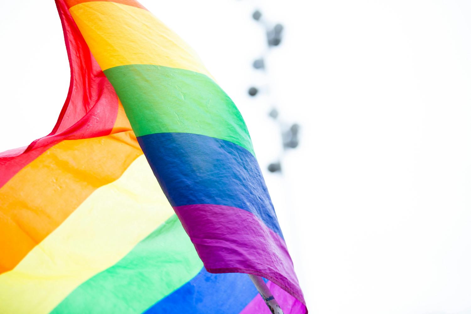 A vibrant rainbow Pride flag, representing LGBTQ+ pride, waves against a bright, softly blurred background.