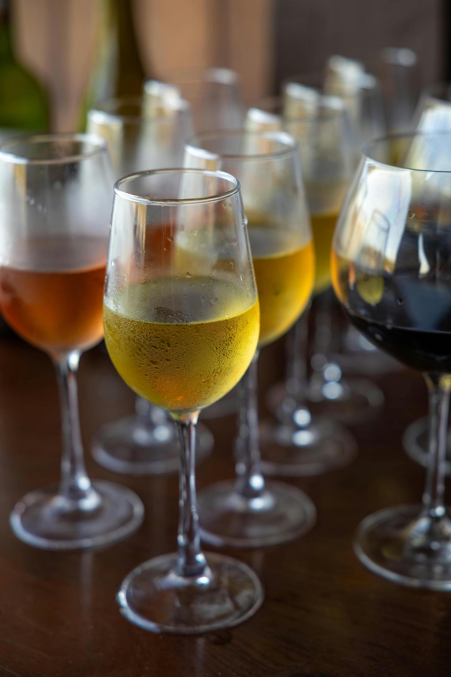 Multiple wine glasses containing rosé, white, and red wines are arranged on a dark wooden table. Condensation beads on the chilled white wine glass in the foreground.