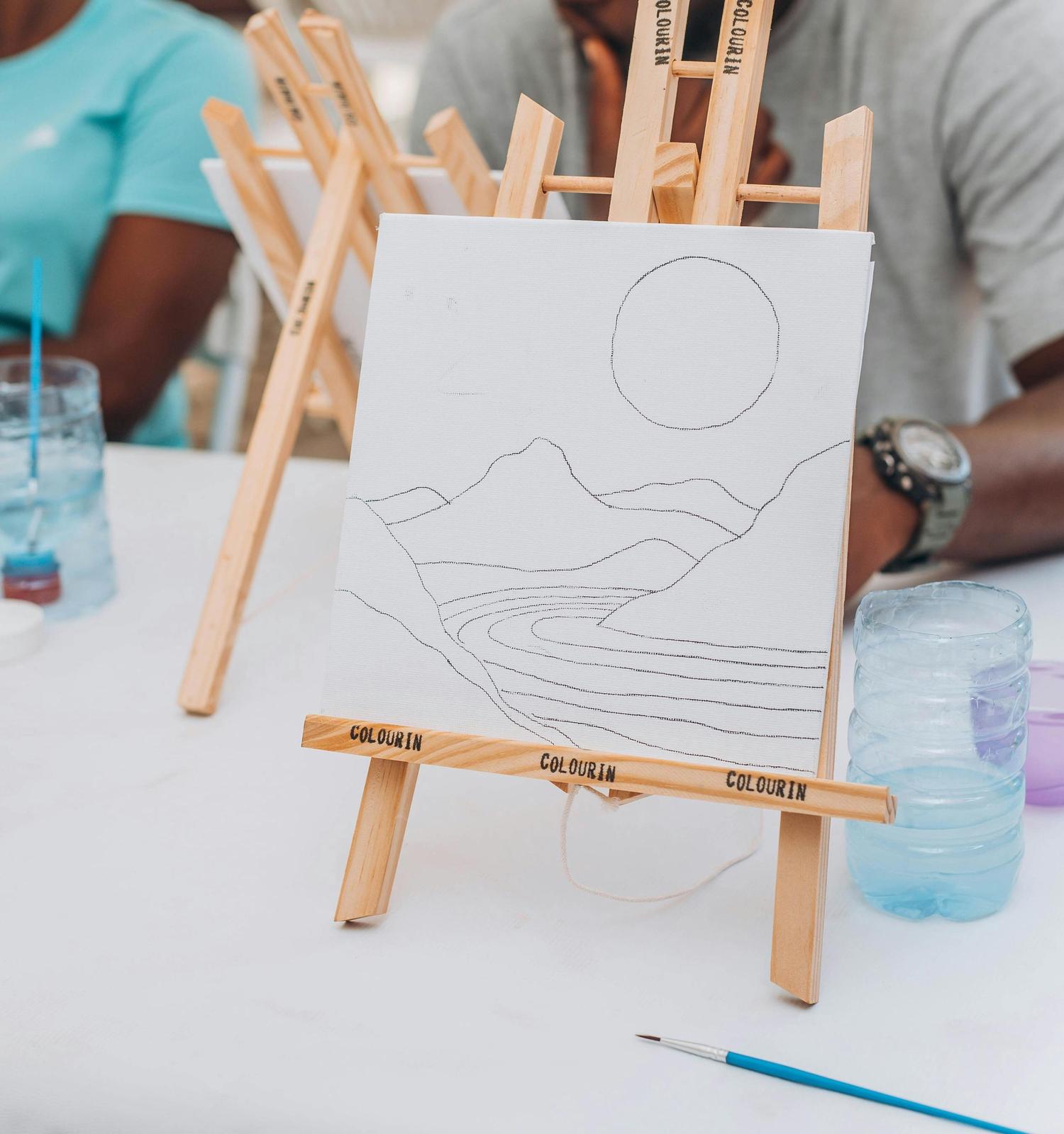 A white canvas on a wooden easel displays a black outline drawing of mountains, a winding river, and a large circle, likely a sun or moon. A blue paintbrush and a water bottle sit on the table, indicating a painting activity in progress.