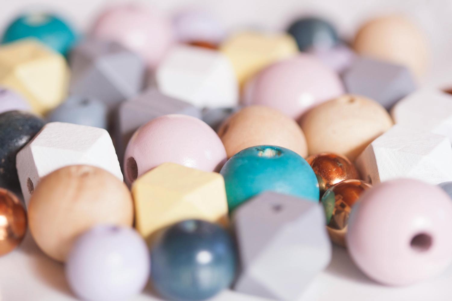 A close-up of a pile of craft beads, featuring various shapes like round and faceted, in a soft pastel palette including pink, cream, yellow, teal, grey, and a few shiny copper ones.