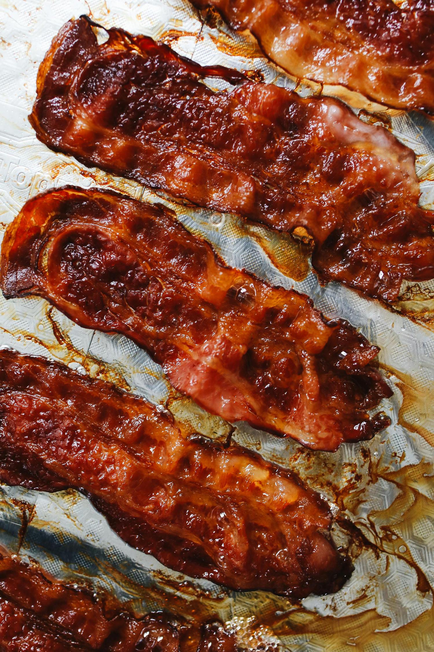 Several strips of glossy, reddish-brown bacon are arranged on a textured baking sheet, glistening with rendered fat.