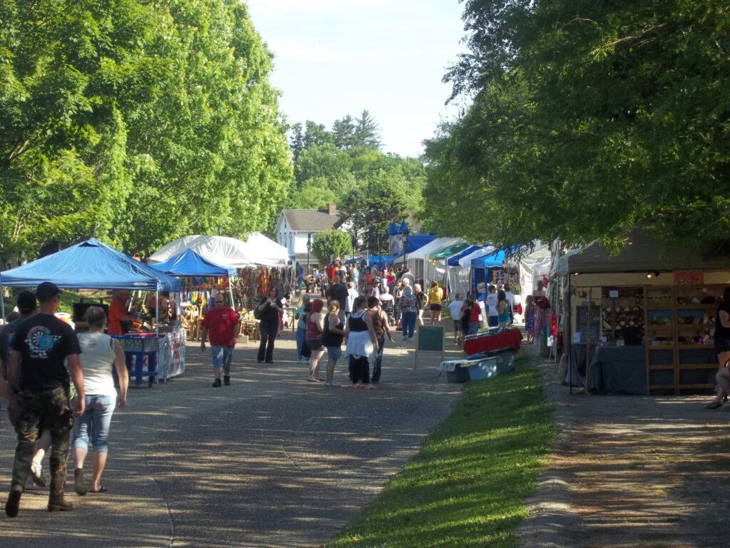 A bustling outdoor market fills a tree-lined path, with people browsing numerous vendor tents set up on both sides.