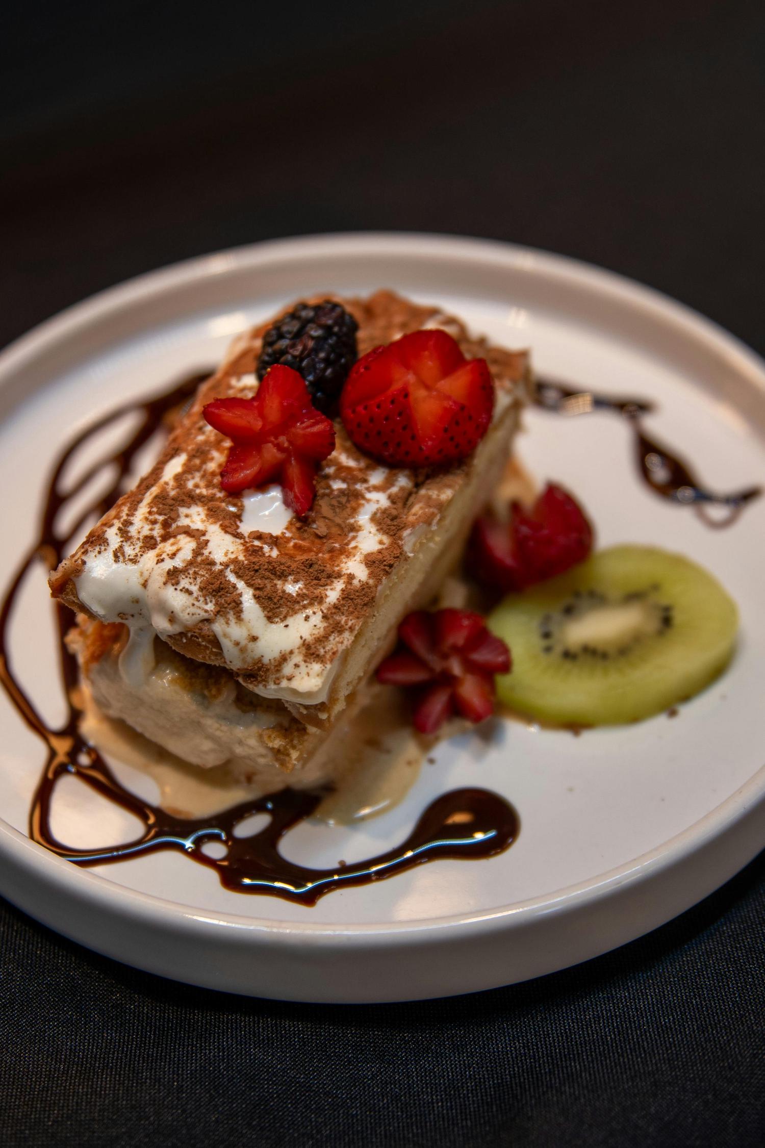 A layered dessert topped with cream, cocoa powder, and garnished with a blackberry, sliced strawberries, and a kiwi slice. Chocolate sauce is drizzled artistically on the white plate around the dessert.
