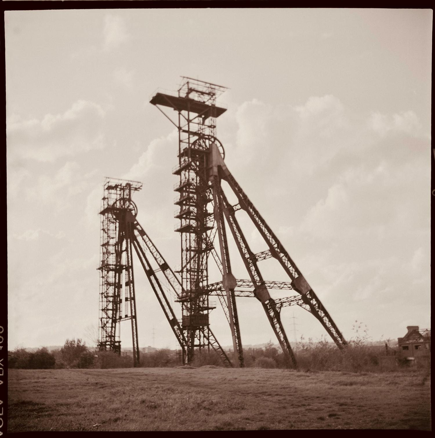 Two towering, skeletal metal structures, resembling old mine headframes, stand in a wide grassy field under a cloudy sky, captured in a sepia-toned photograph.
