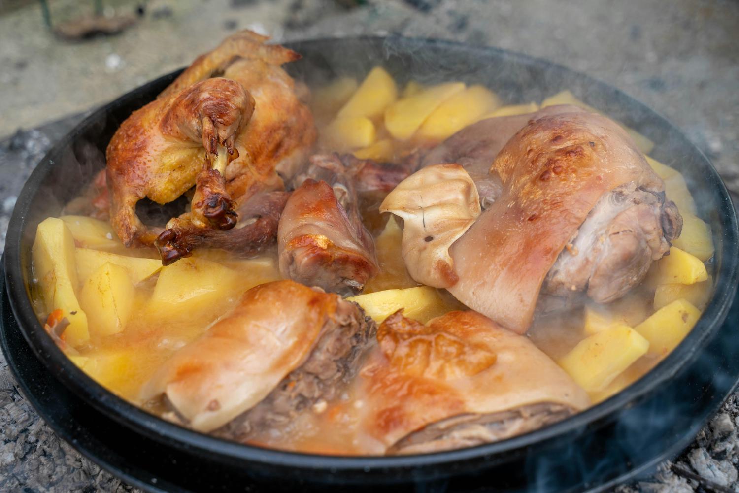 A large cast iron pot holds a whole roasted chicken, pork pieces, and chunky potatoes simmering in a golden broth, with steam rising. The pot rests on a bed of ashes, indicating outdoor cooking.