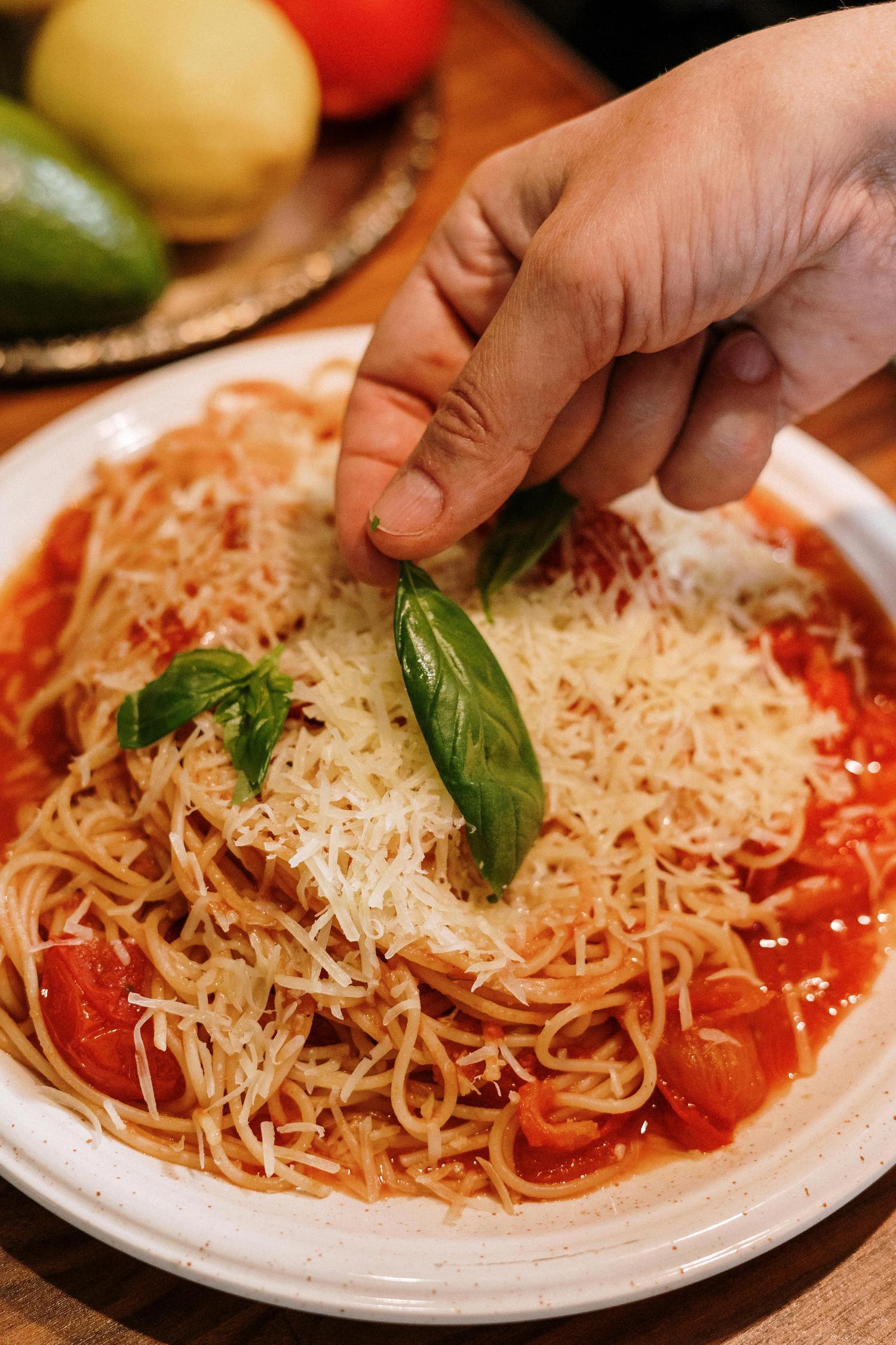A hand adds fresh basil leaves to a plate of spaghetti topped with tomato sauce and grated cheese, with blurred fruits visible in the background.