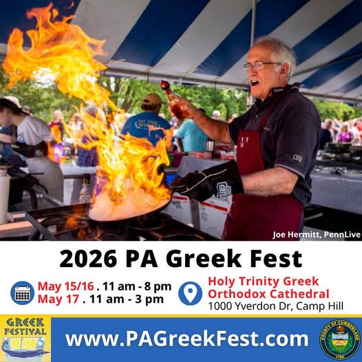 A chef wearing an apron and grill mitt enthusiastically flambés food, sending large flames upward from a pan, at an outdoor Greek festival under a striped tent.